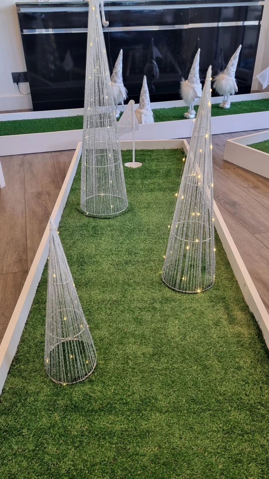 Decorative holiday display with white wire cone trees with lights on green artificial grass and small white gnome statues in the background.