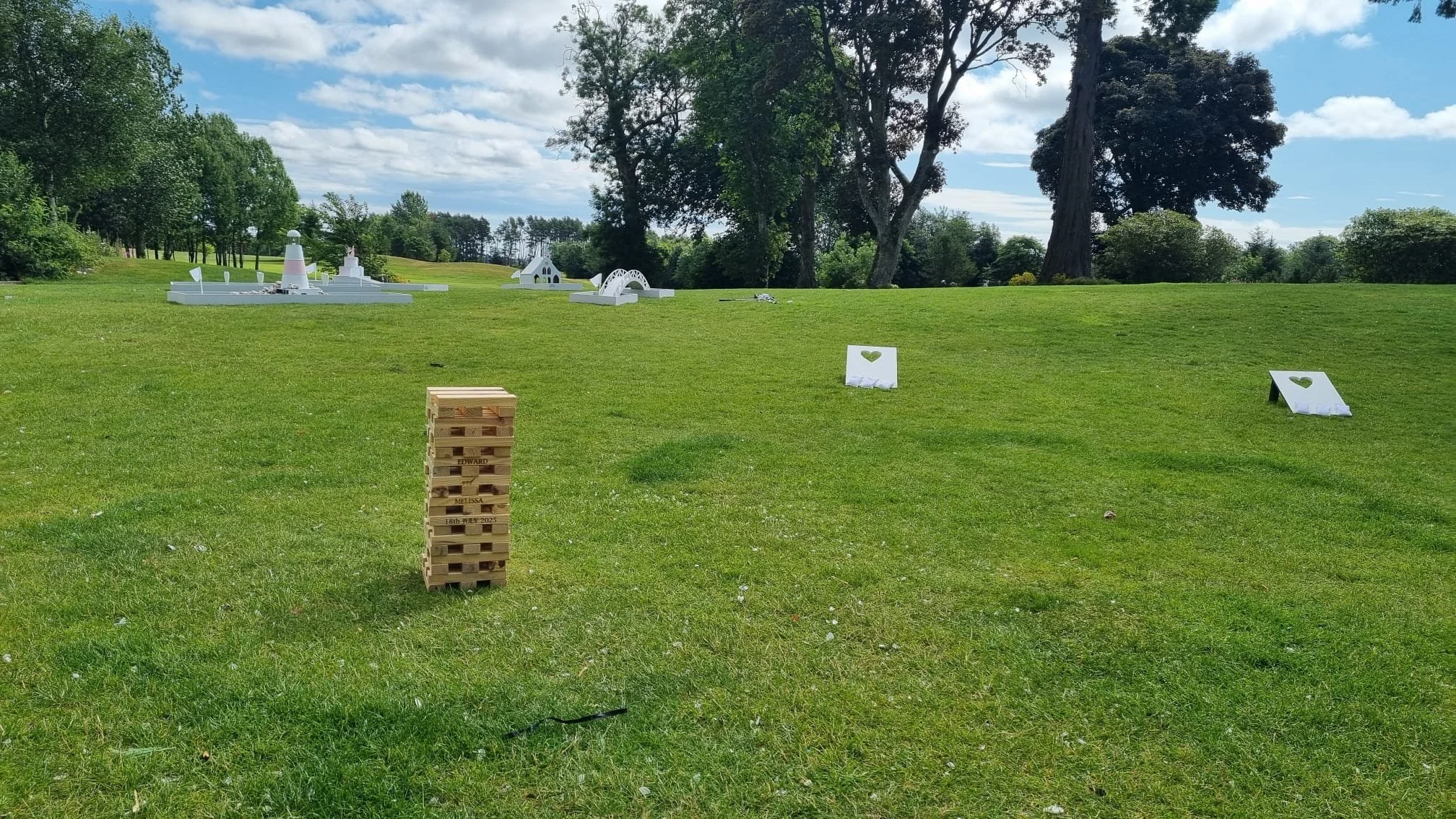 Green grassy field with white decorative structures in the distance, including a lighthouse, bridges, and small buildings. There are two white signs with green hearts and a wooden Jenga tower in the foreground. Tall trees and a partly cloudy sky are 