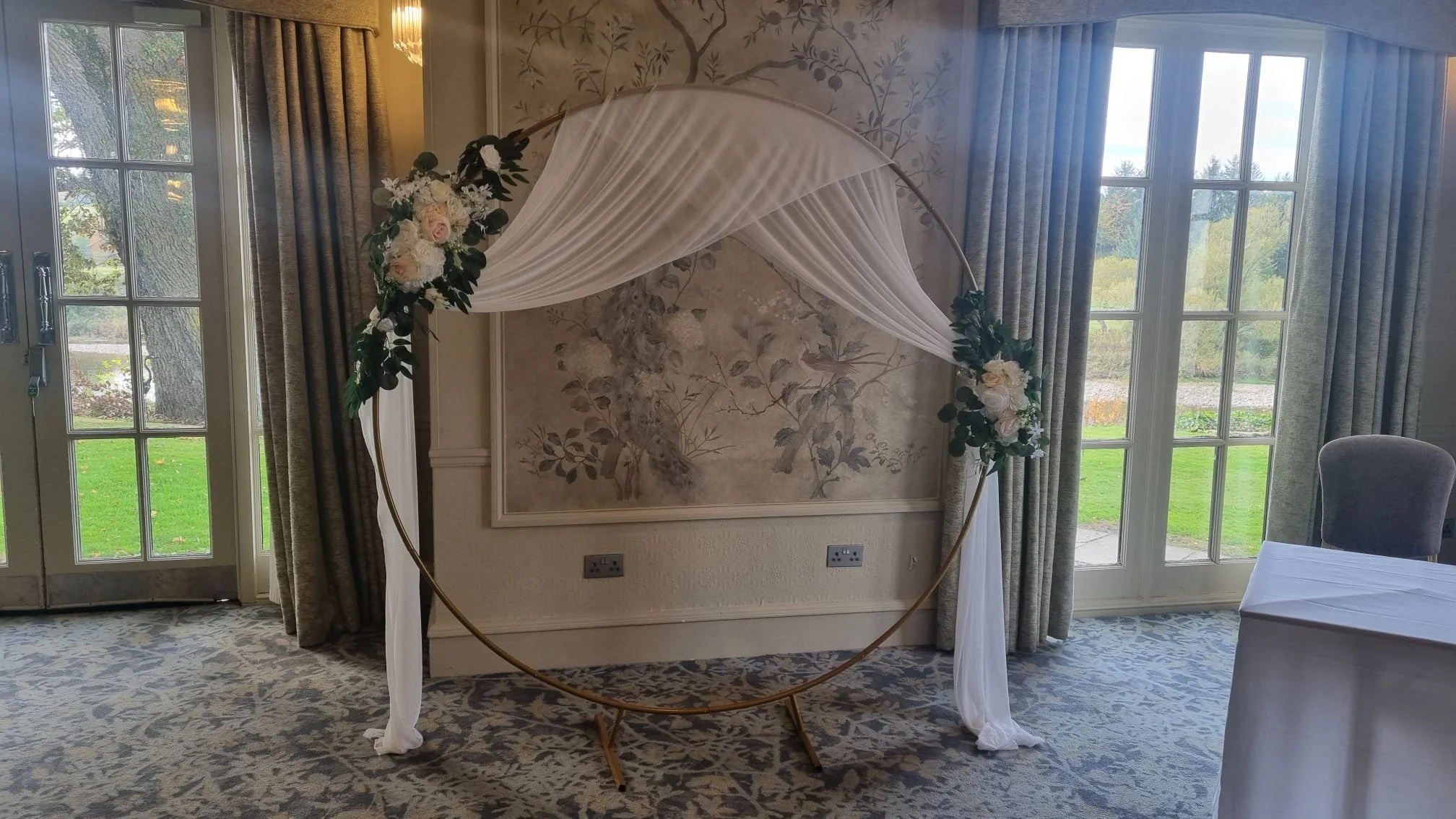 A floral wedding arch with a circular gold frame, decorated with white fabrics and white and cream flowers with greenery, set indoors with large windows and curtains.