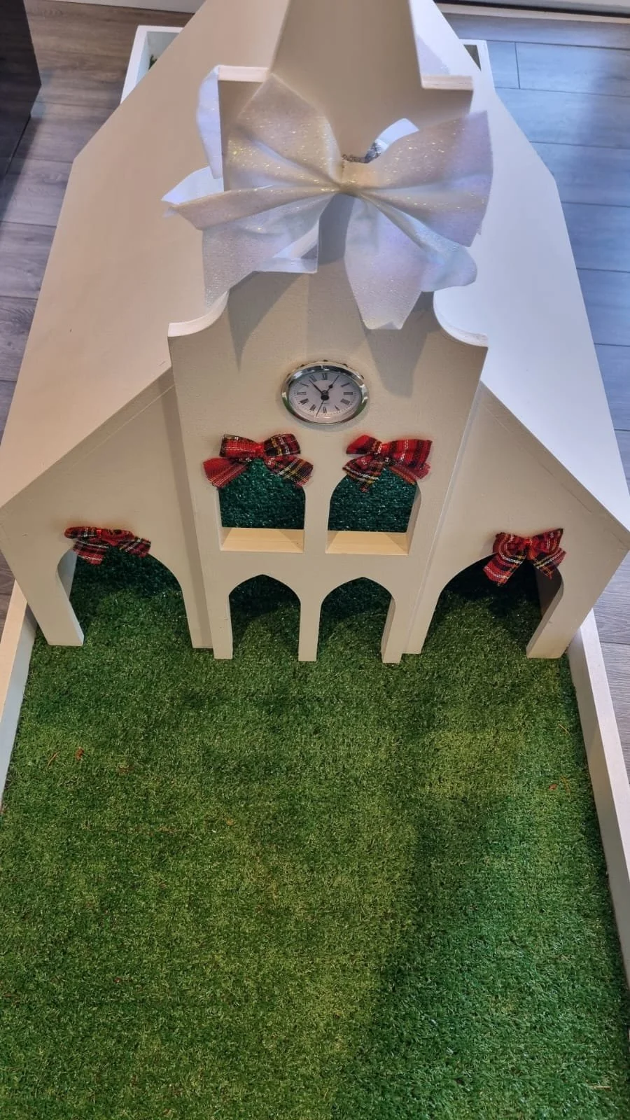 A decorative white wooden house-shaped frame with a large white bow on top, a small clock, and three small Christmas stockings with red plaid bows, all on a patch of green artificial grass.