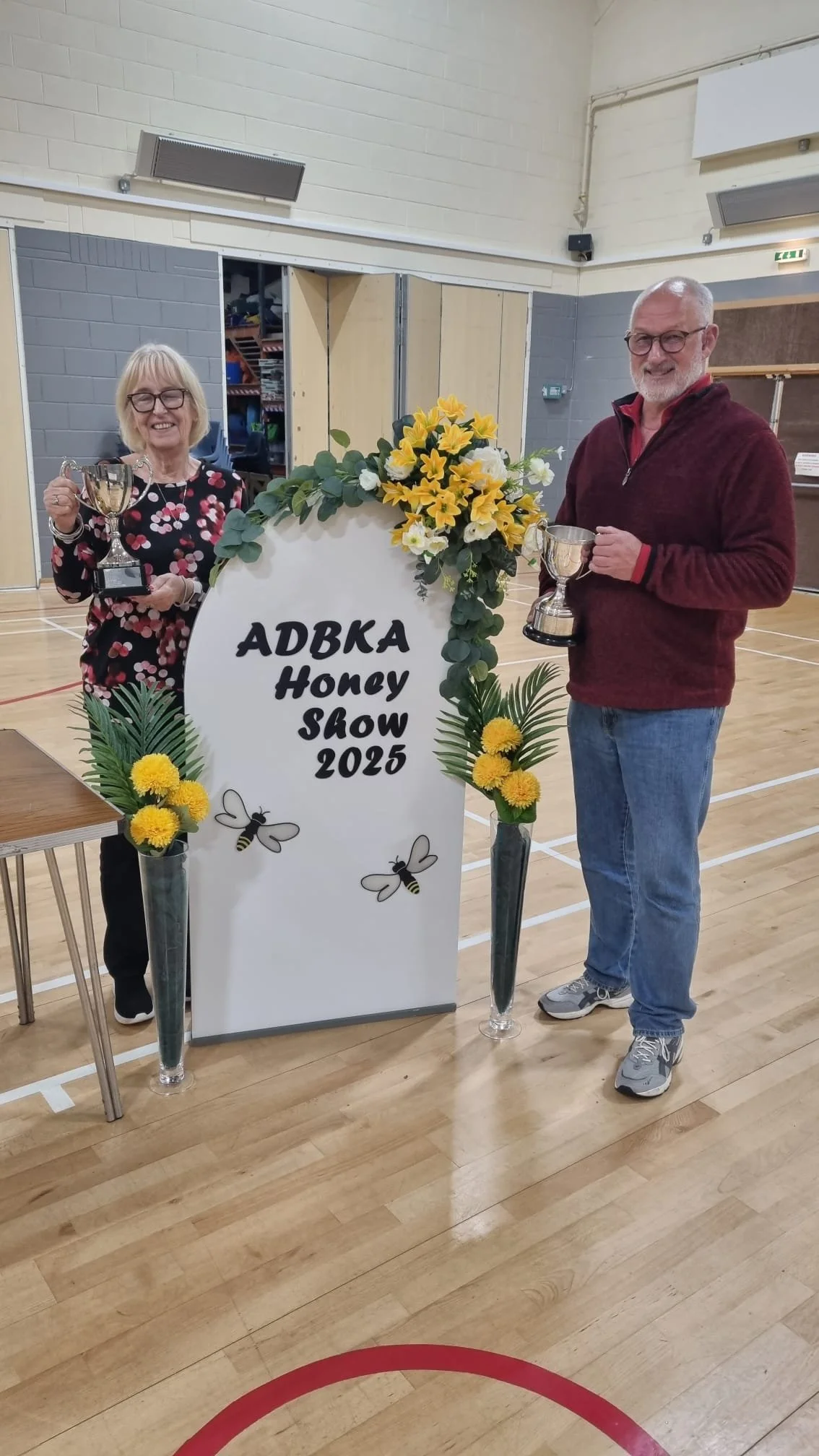 Two people standing on a gymnasium floor holding trophies, celebrating at the ADBKA Honey Show 2023, with a large floral display and a sign in the middle.