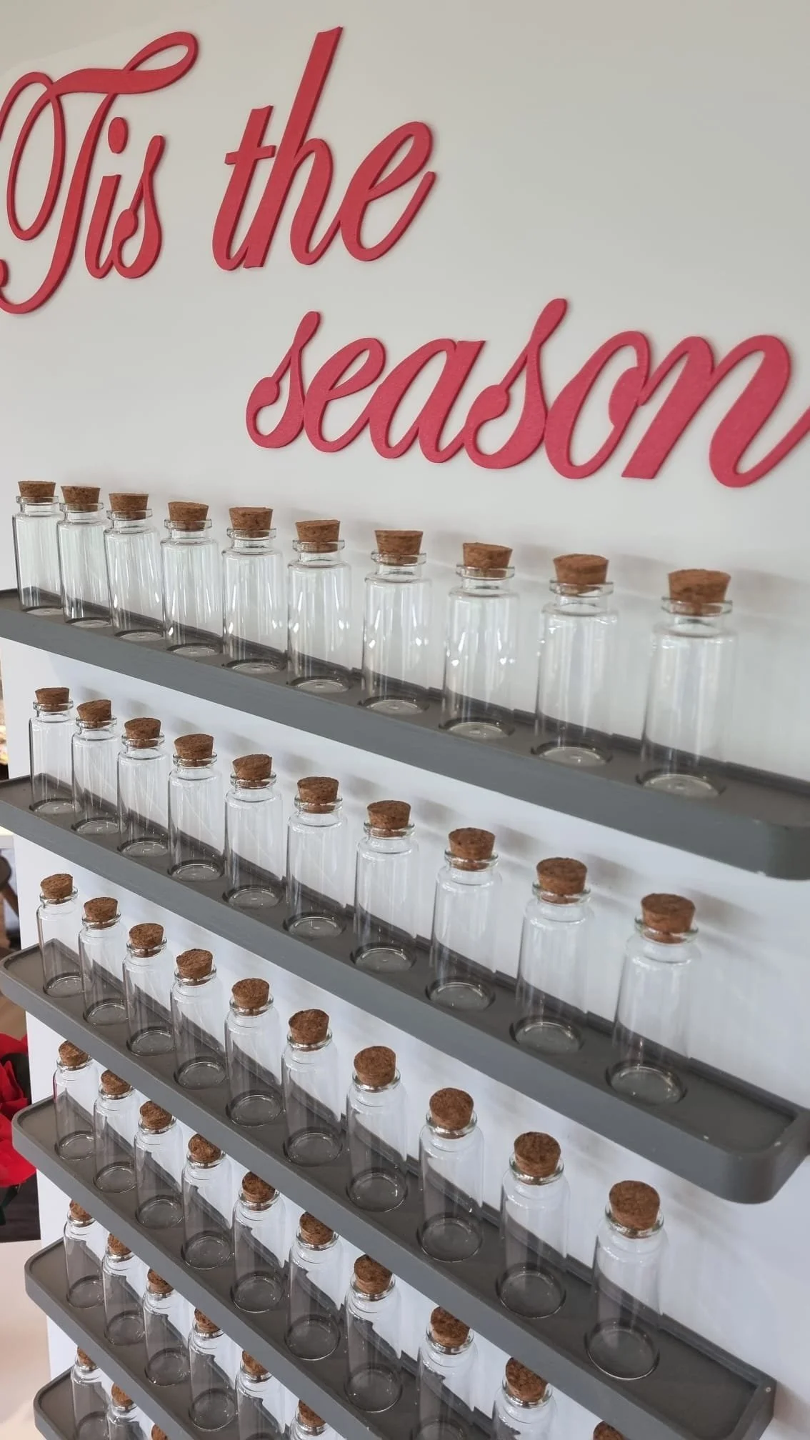 Empty glass jars with cork stoppers arranged on gray shelves against a white wall with pink text that reads 'Tis the season'.