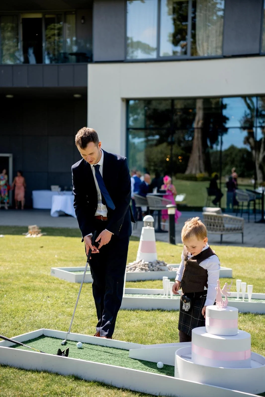 A man in a suit and a young boy dressed in formal attire playing mini golf outdoors near a modern building with large windows, with other people in the background at a social event.