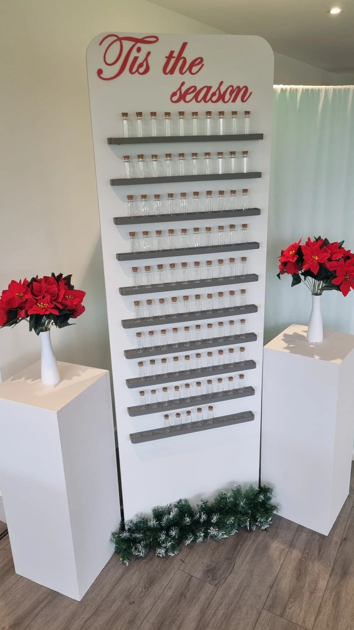 Display stand with glass jars with cork lids, decorated for Christmas, with red poinsettia flowers on white pedestals.