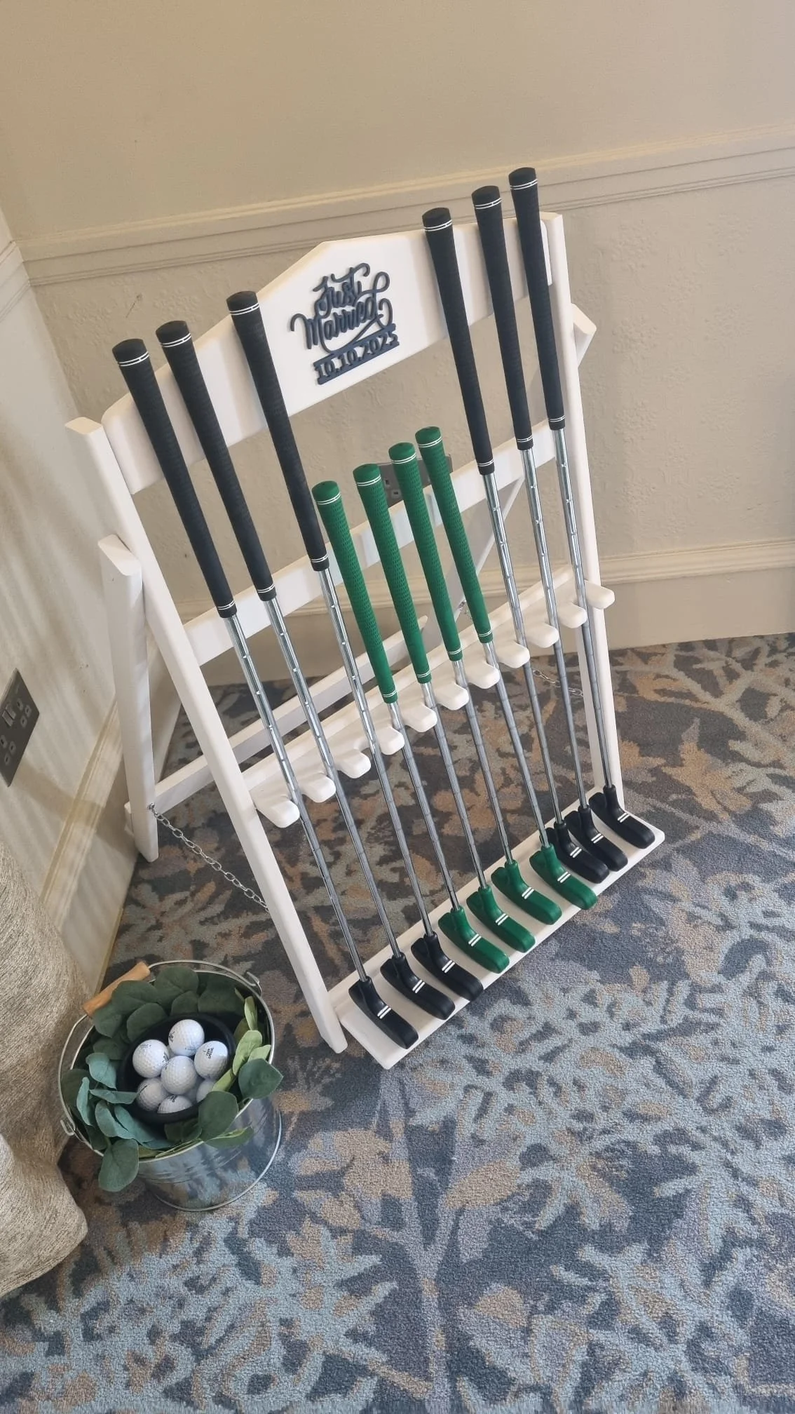 A white golf club display stand with ten golf clubs, five with black grips and five with green grips, positioned upright. The stand has a decorative sign at the top that says "Just Married" with the date "10.10.2023." Next to the stand, there is a ba