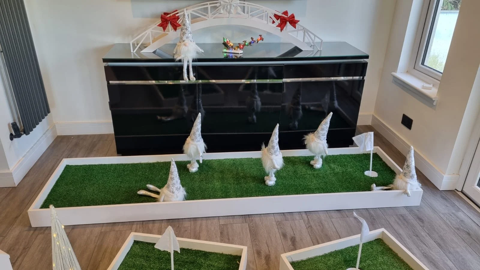 Indoor Christmas decoration with a black cabinet, a white box with green grass and decorated gnome figurines with tall silver hats, pennants, and reindeer antler ornaments.