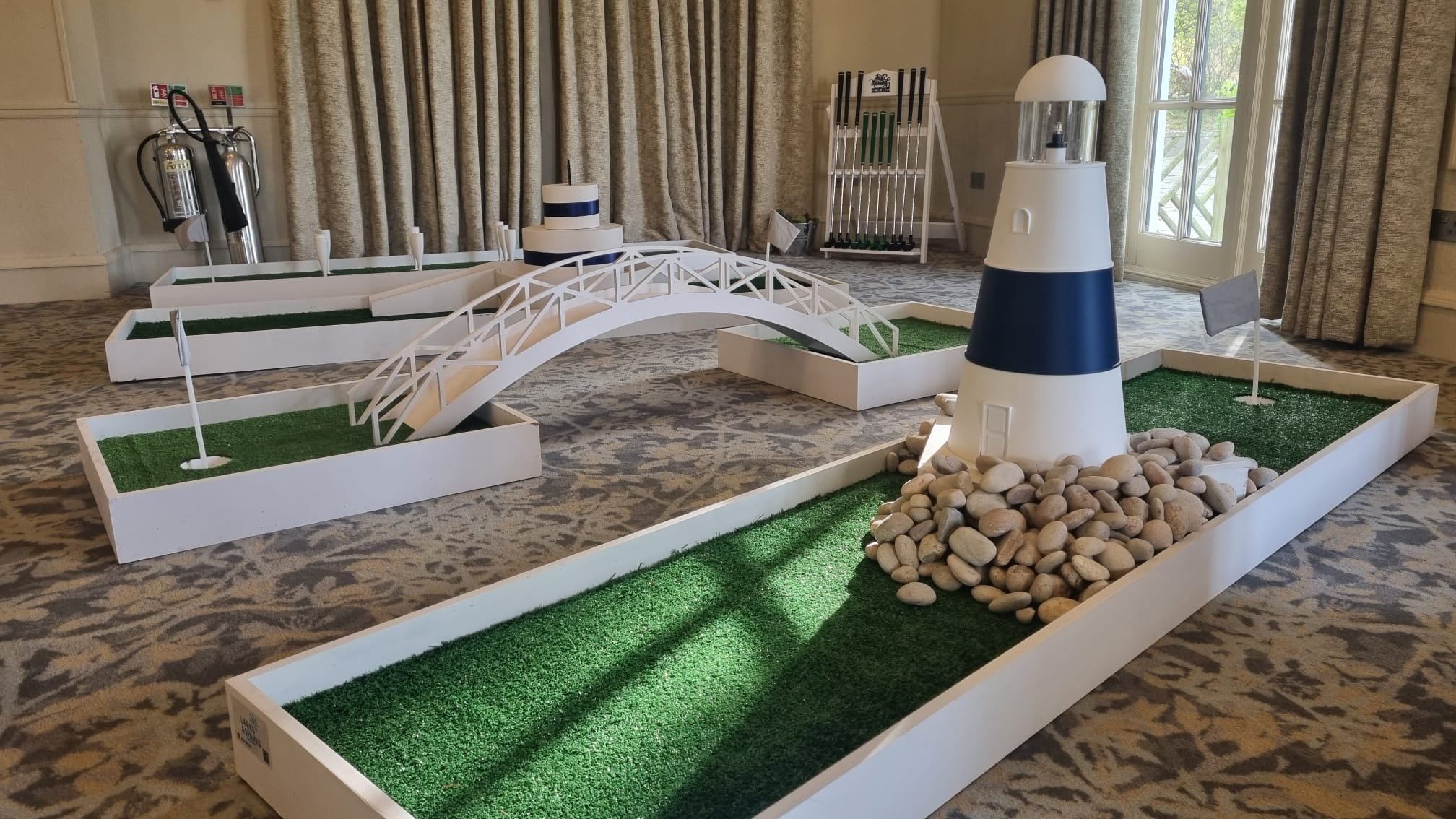 Miniature golf course setup with a lighthouse obstacle, small flags, a pebbled base, and a white bridge on artificial turf in an indoor space.