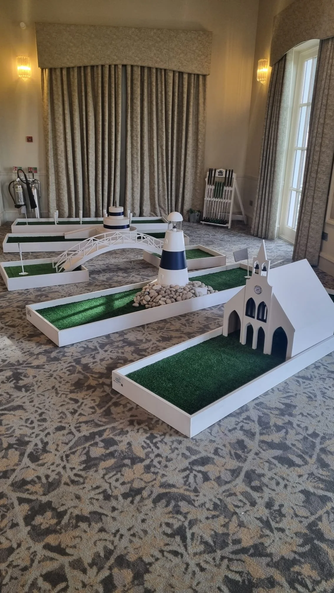 Miniature golf course display with a small white church, a lighthouse, a bridge, and scattered pebbles, set indoors on a patterned carpet.
