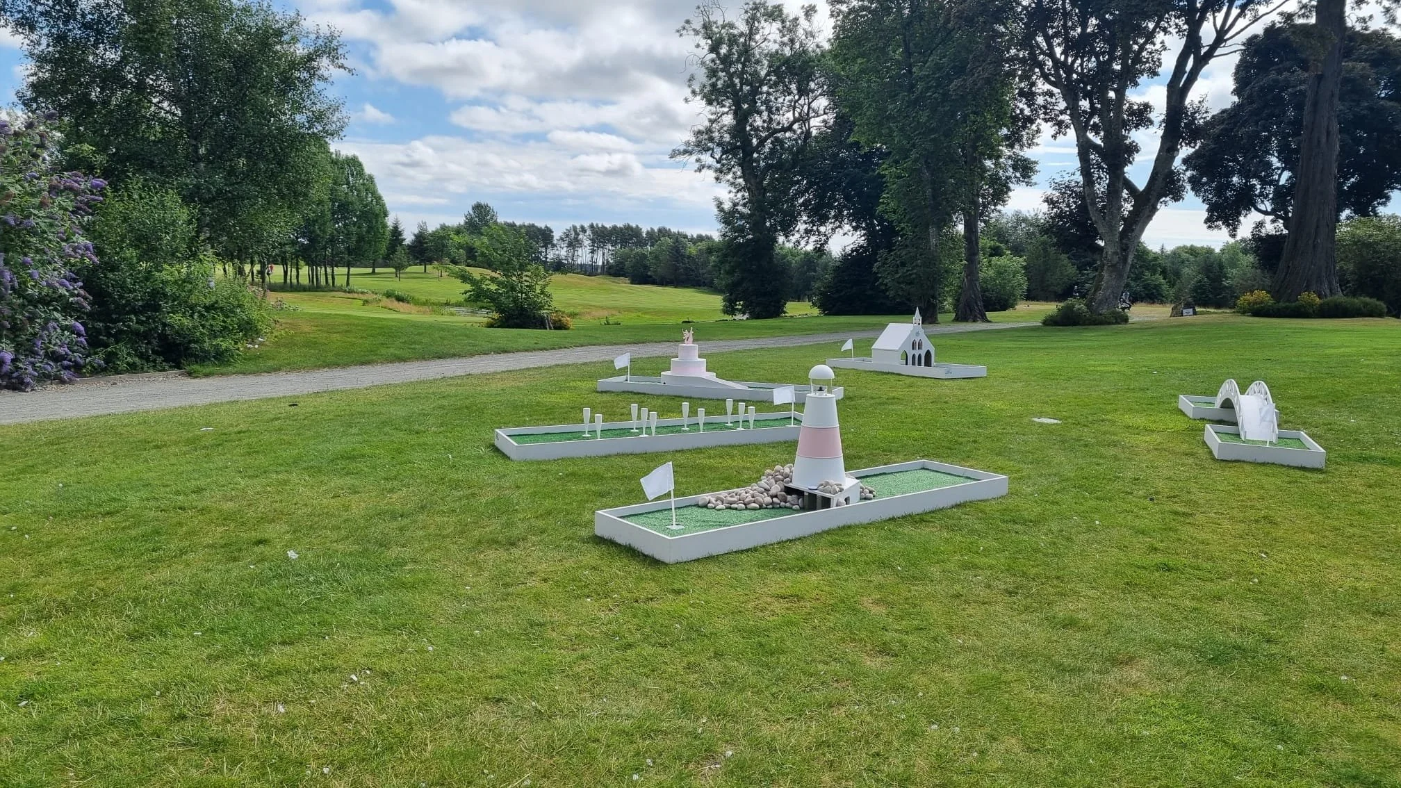 Miniature golf course with small themed structures, including a lighthouse, church, and bridge, set on a well-maintained grassy area with trees and a cloudy sky in the background.