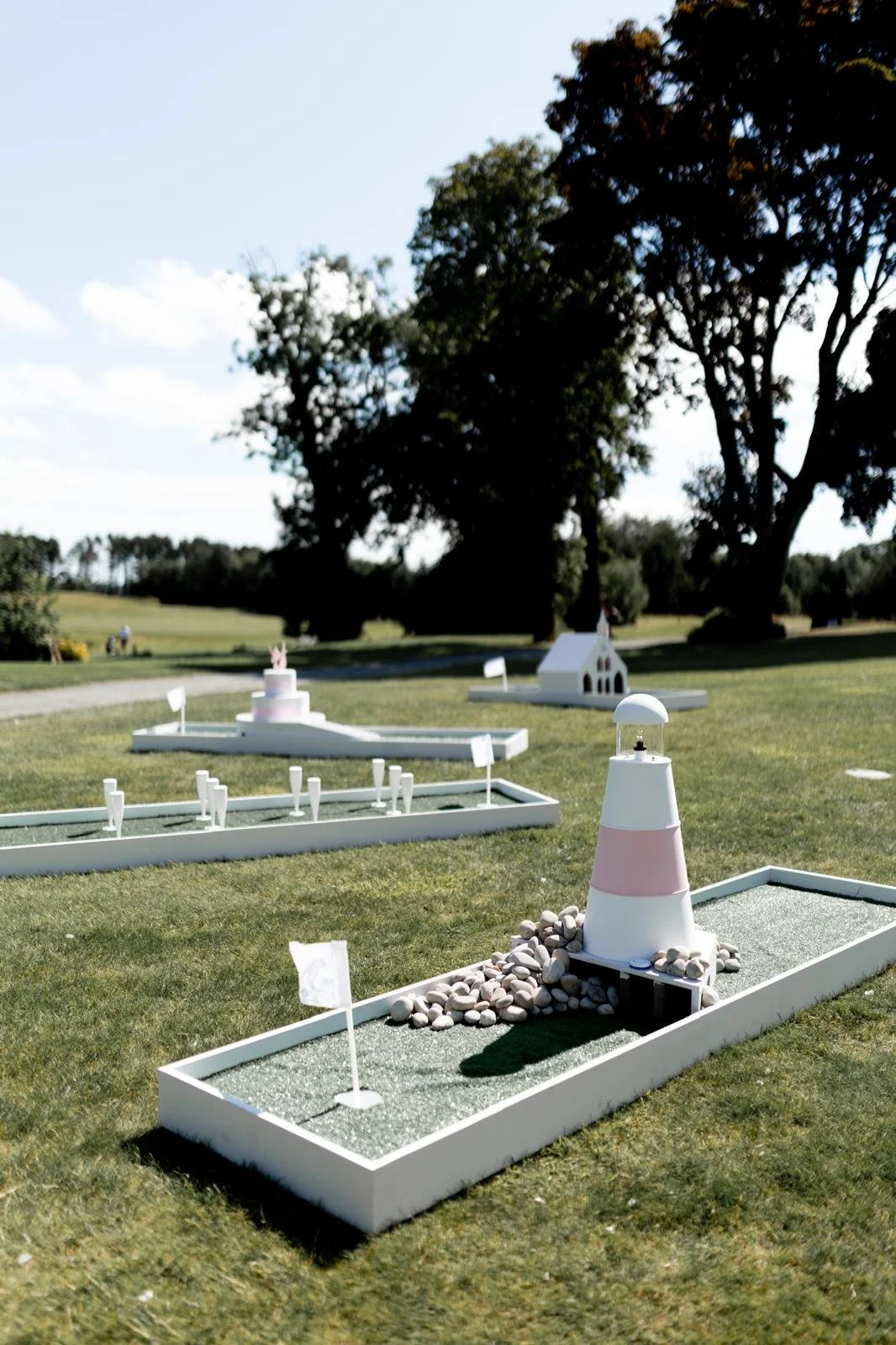 Miniature golf course with lighthouse-themed holes, green artificial turf, rocks, and flags under a partly cloudy sky with trees in the background.