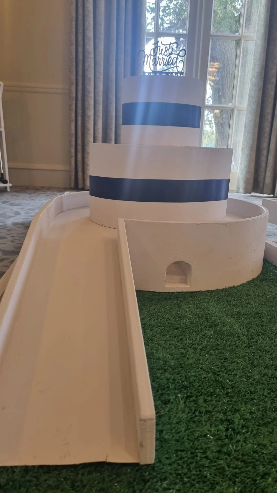 Miniature model of a wedding cake with a 'Just Married' topper, placed on a wooden surface with artificial green grass and a slide in the foreground, inside a room with windows and curtains.