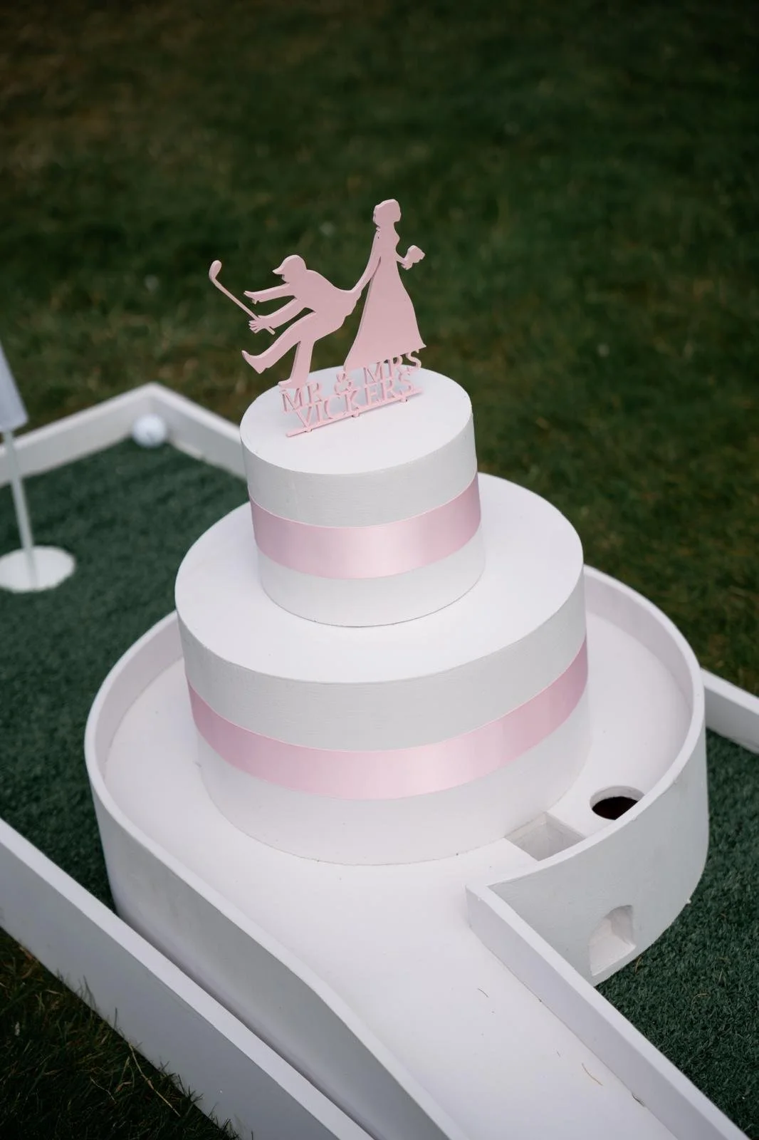 A three-tiered white cake with pink ribbons, topped with a cake topper that says 'Mr & Mrs Vickners', showing a silhouette of a woman holding a golf club and a man in a golf pose.