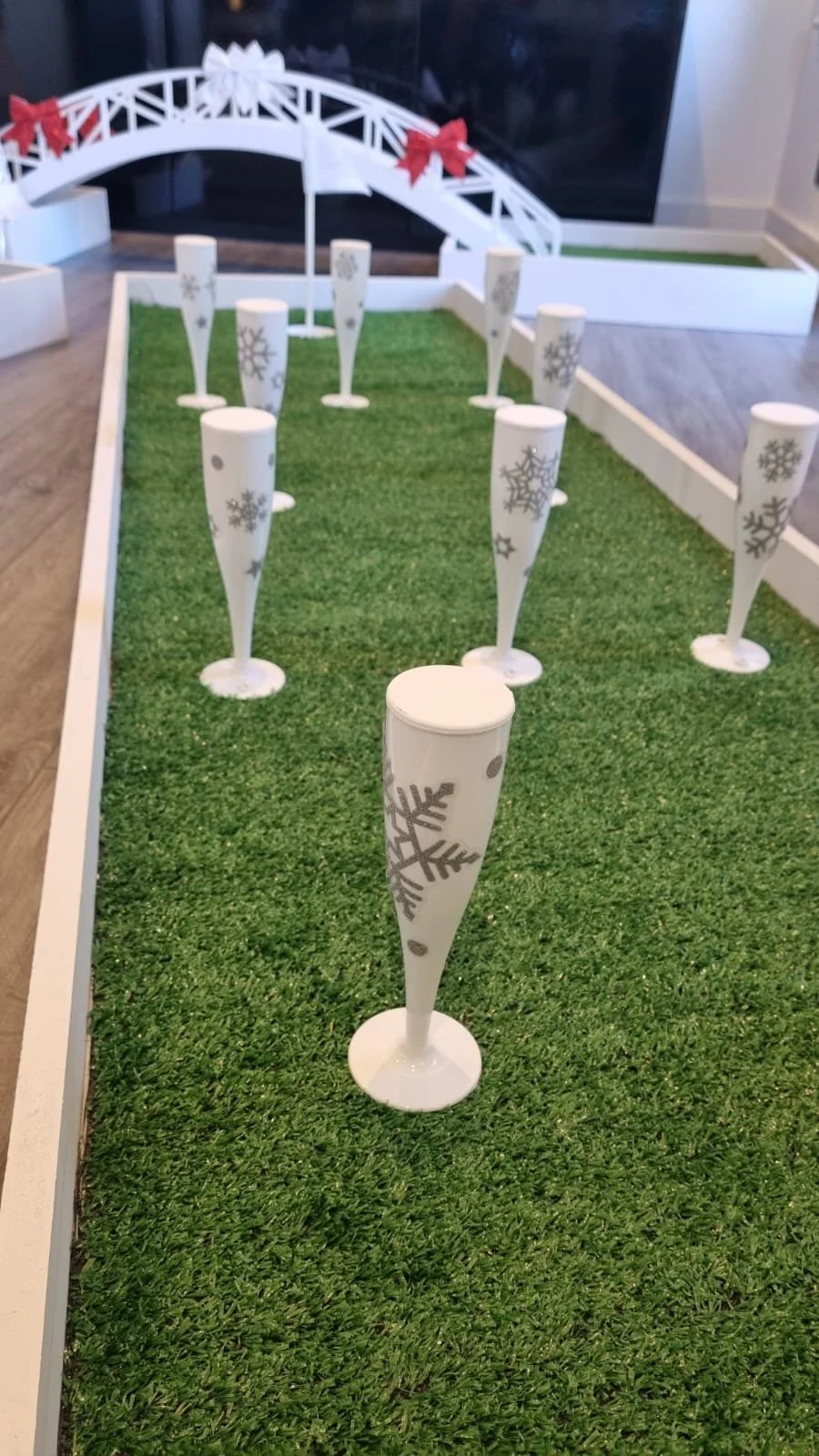 Miniature golf course with white cones decorated with snowflakes, set up on artificial green grass, with a white bridge and red bows in the background.