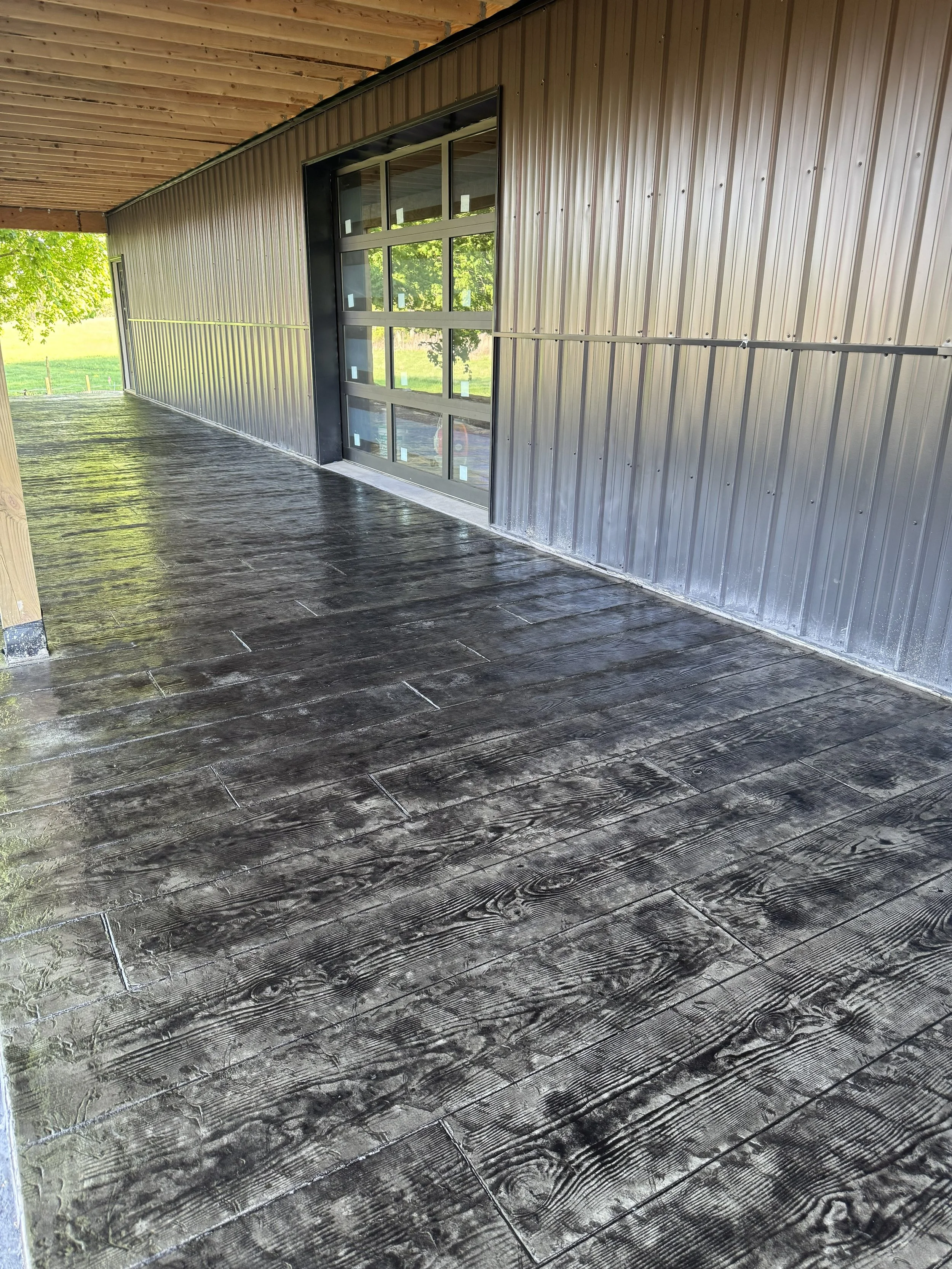 Interior of a covered porch with a new concrete floor, metal siding wall, and large glass garage door, outdoors with trees and green grass visible.