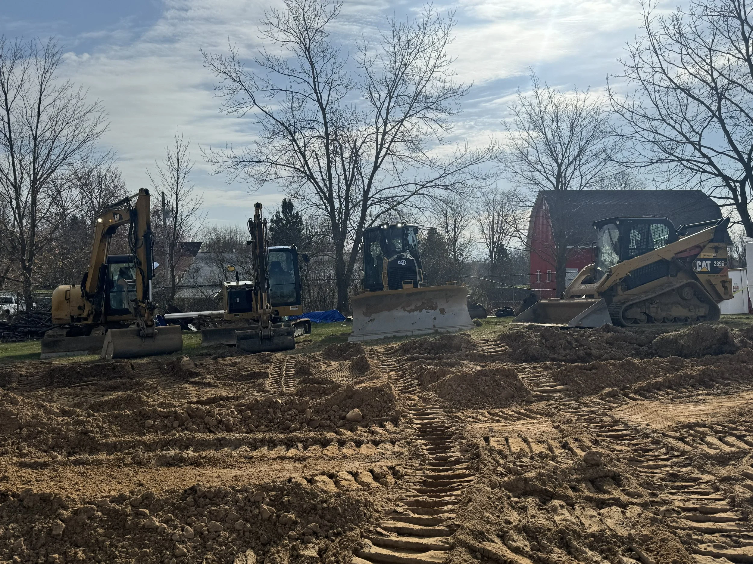 Four pieces of construction equipment, including two mini excavators, a bulldozer, and a tracked loader, are parked on a dirt construction site with tire tracks and cleared ground, with leafless trees and a red barn in the background under a partly cloudy sky.