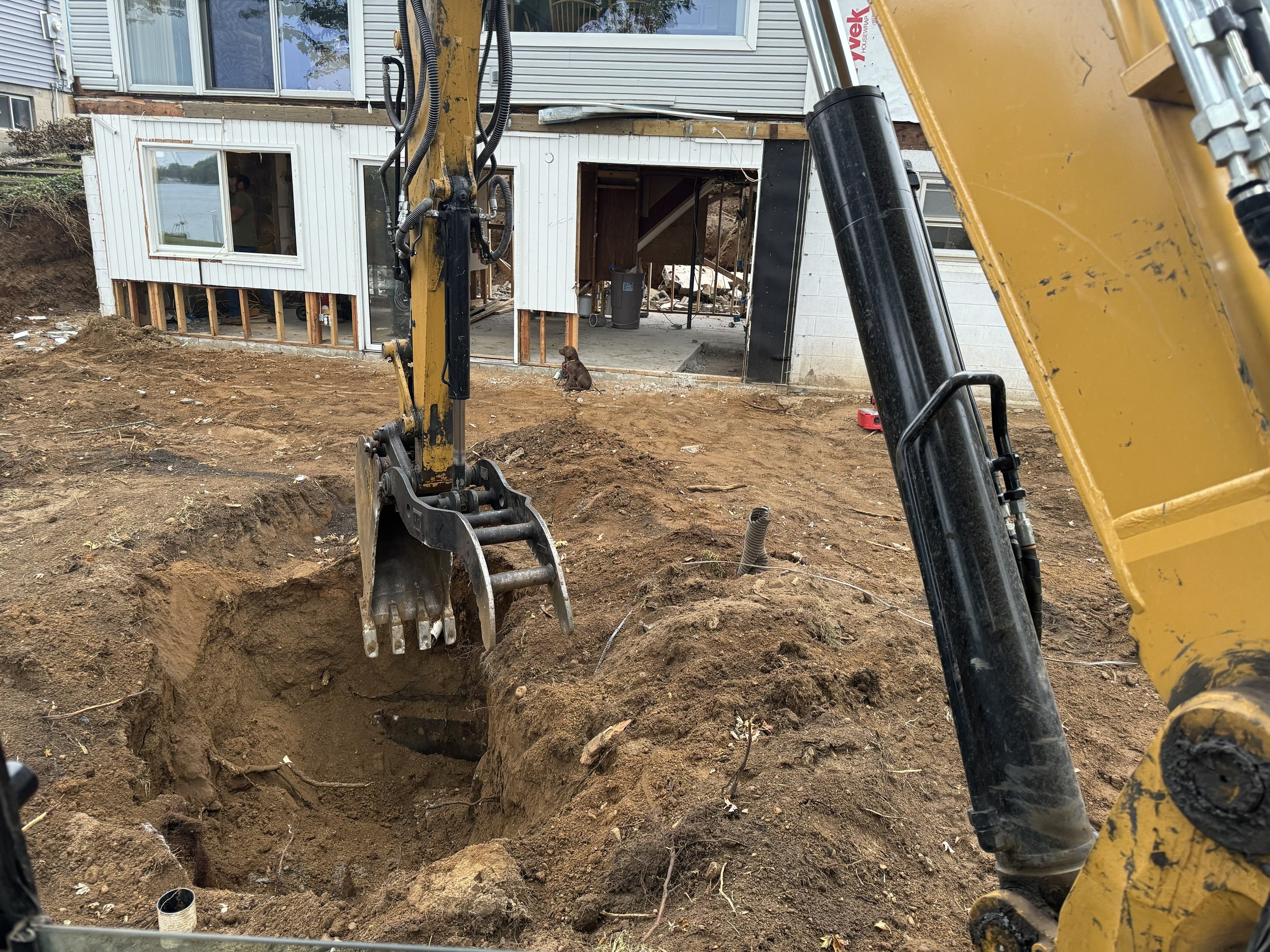 Construction site with excavator digging a hole in front of a house, showing exposed foundation and open basement area.