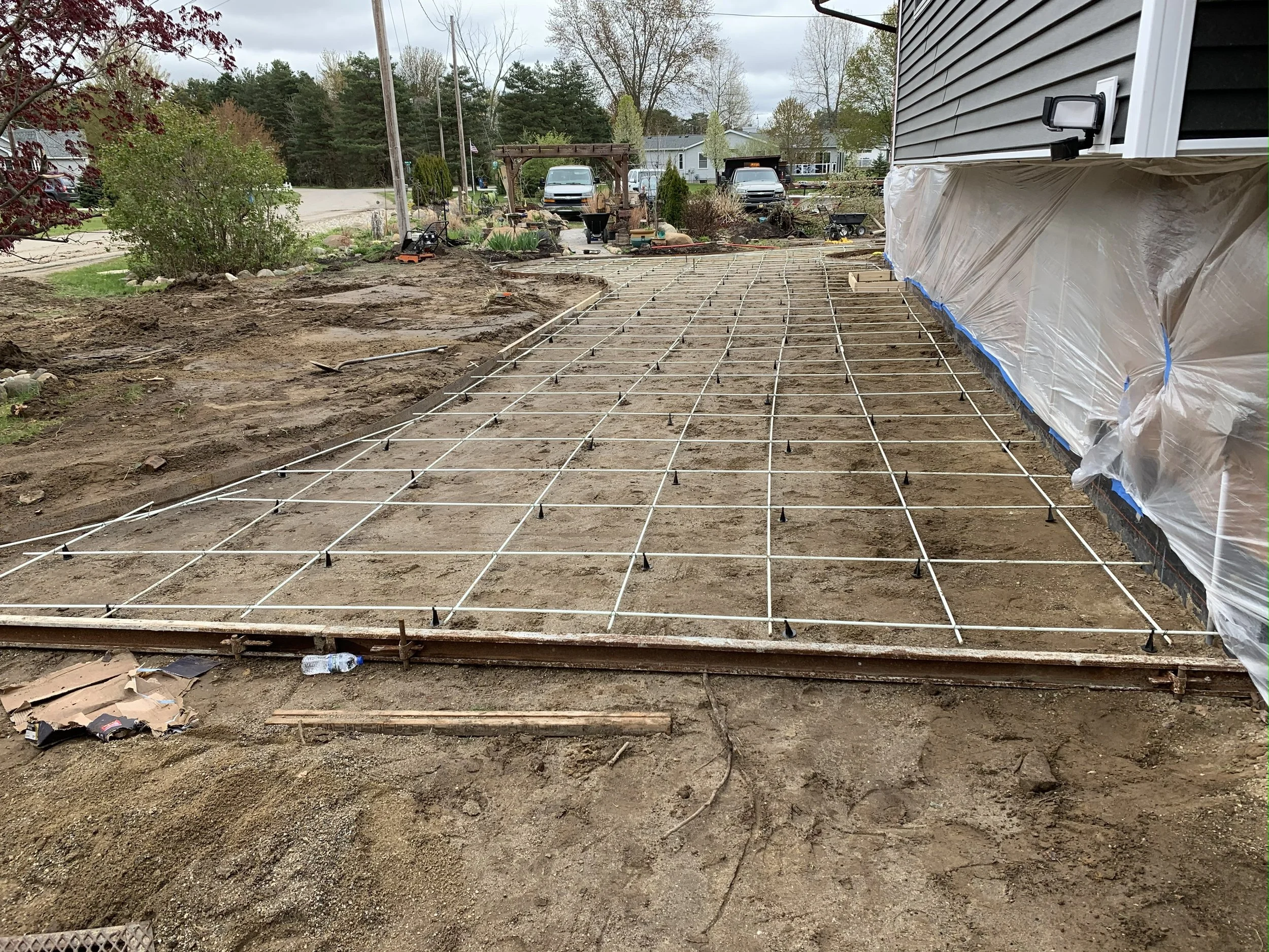 Construction site with a grid of rebar for a concrete slab in front of a house, with plastic sheeting covering part of the house exterior.