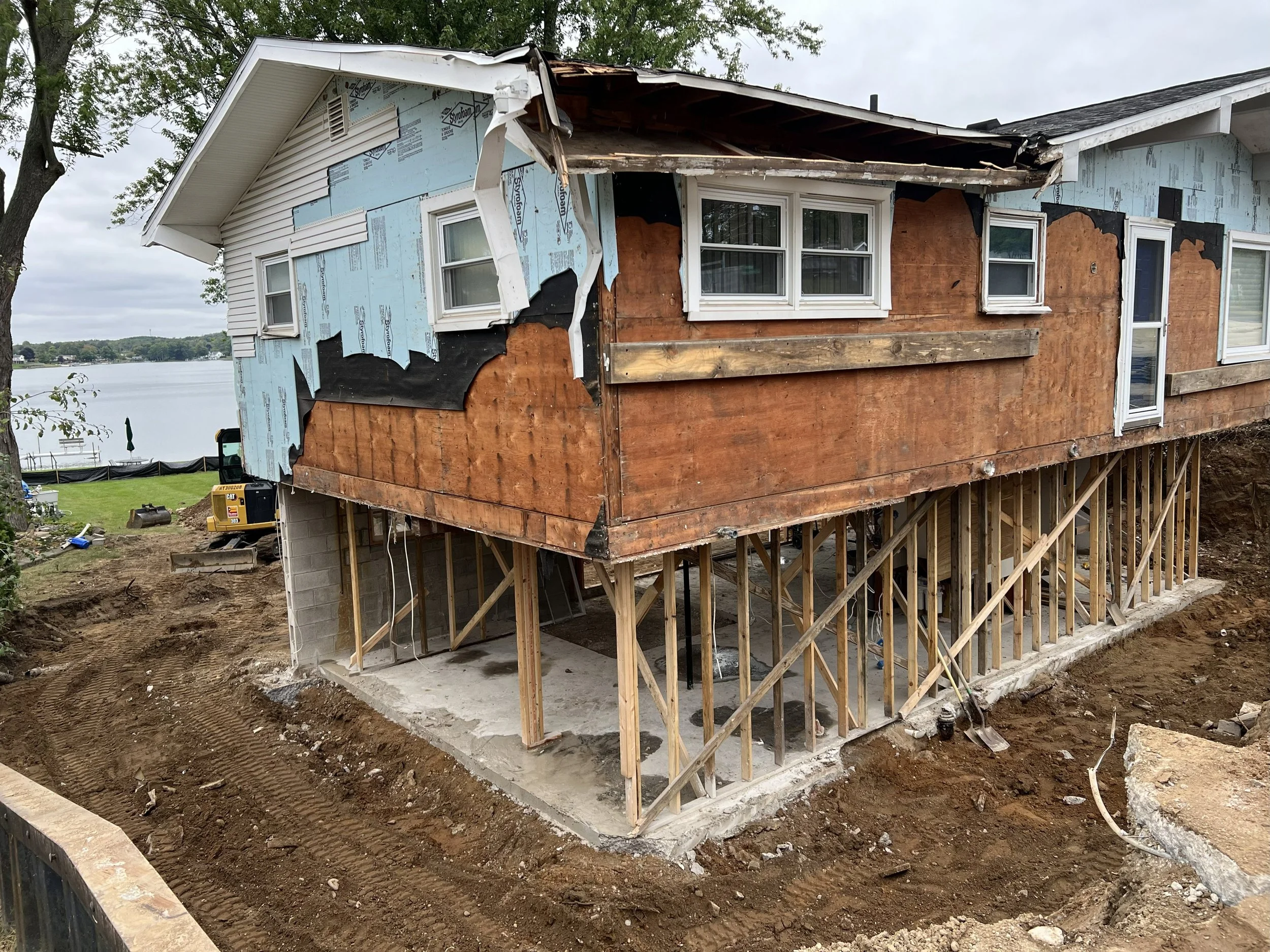House under construction extending over a body of water with visible window frames and partially finished exterior walls, supported by wooden scaffolding.