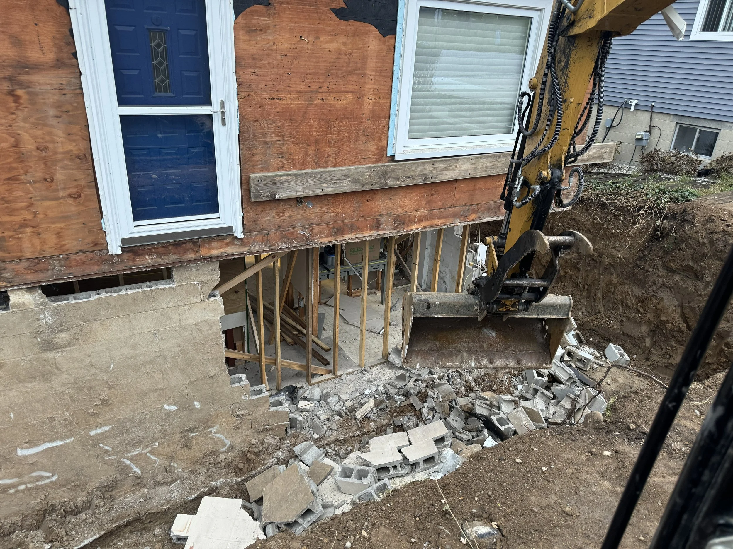 Construction site showing a bulldozer bucket with an excavator at a house undergoing renovation. The house has a partially exposed foundation with tracks and cinder blocks on the ground, and the exterior wall is being worked on with plywood and woode