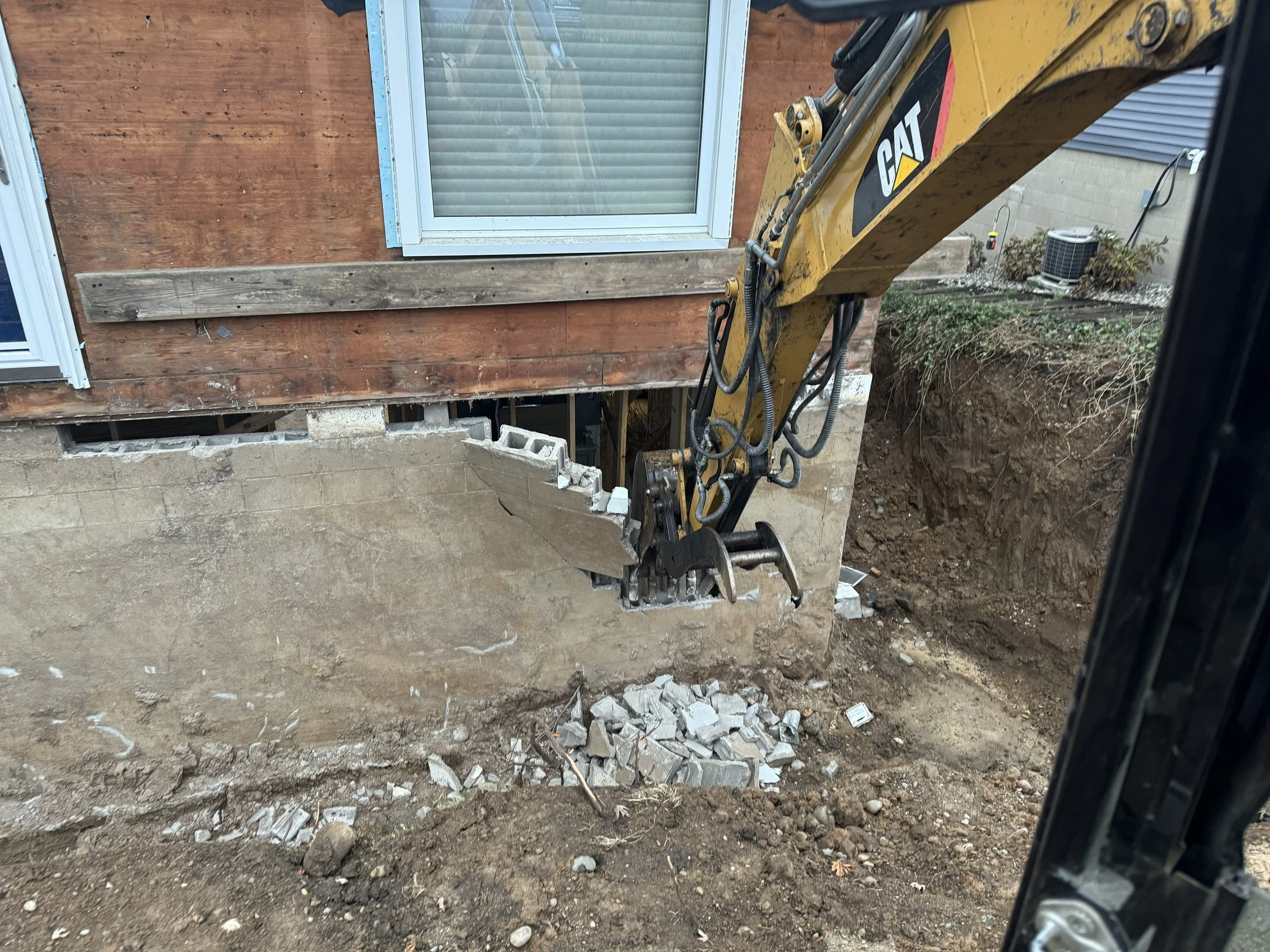 Construction work with a backhoe removing part of a concrete foundation under a house with a wooden wall and windows, and an excavated dirt area nearby.