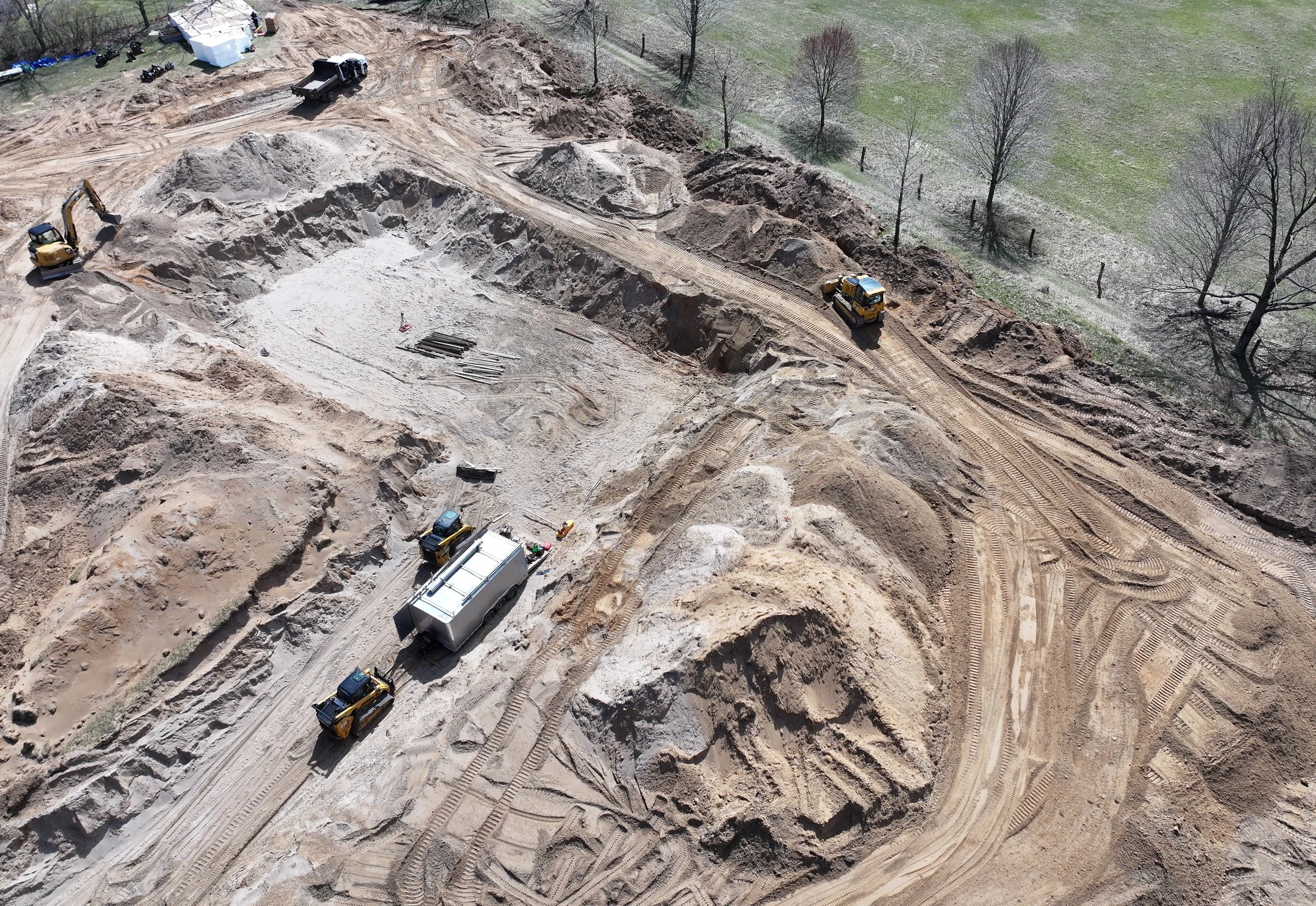 Construction site with excavators and trucks, showing dirt piles and cleared area, near a grassy area with trees.
