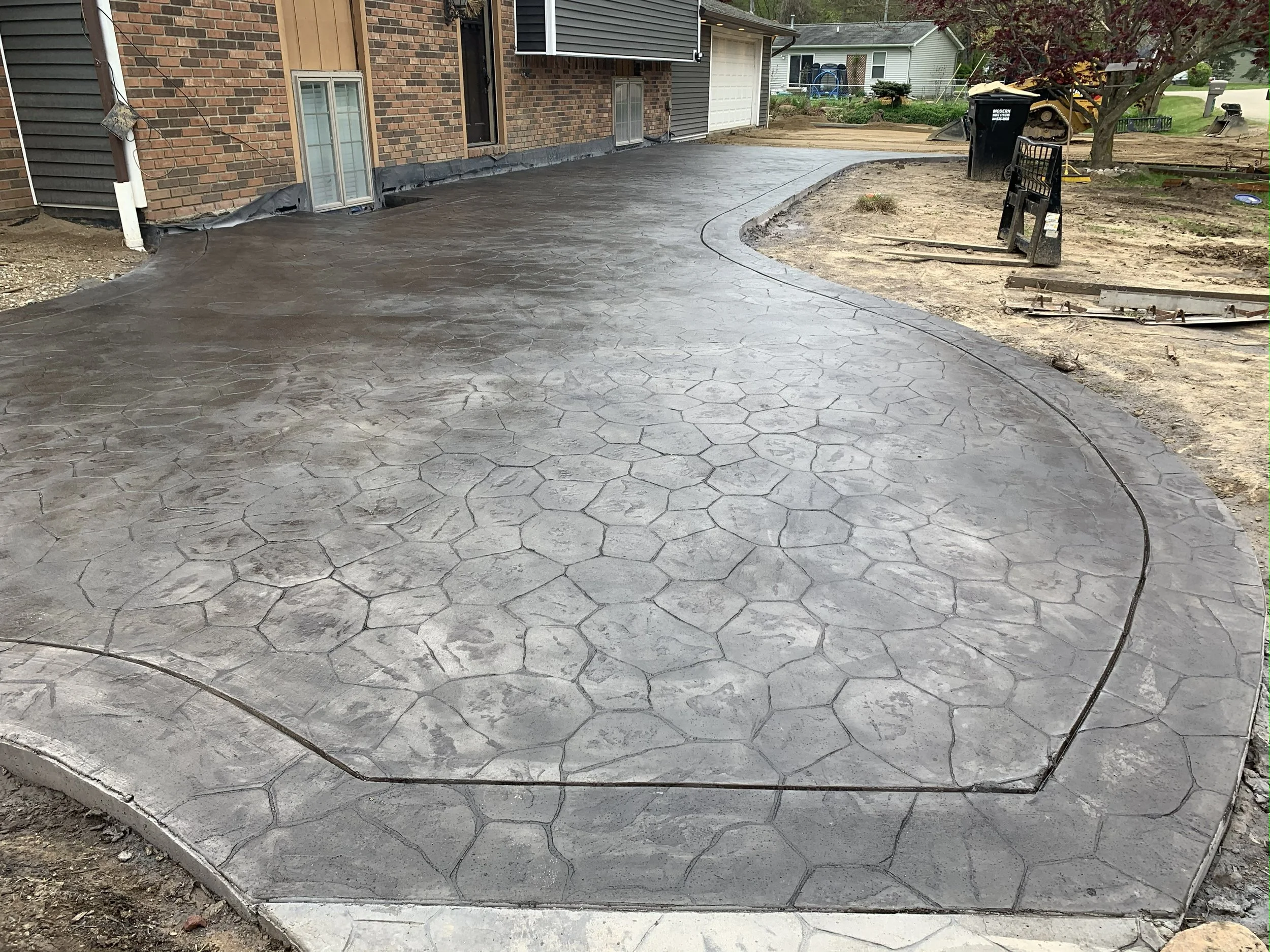 Freshly poured and sealed concrete driveway with a decorative stamped finish, curving around a house under construction.