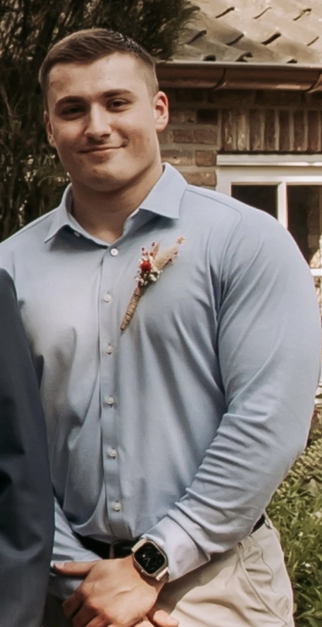 Een jonge man met een lichte blauwe overhemd en een smartwatch, zittend buiten in een tuin, met een bakstenen huis en planten op de achtergrond.