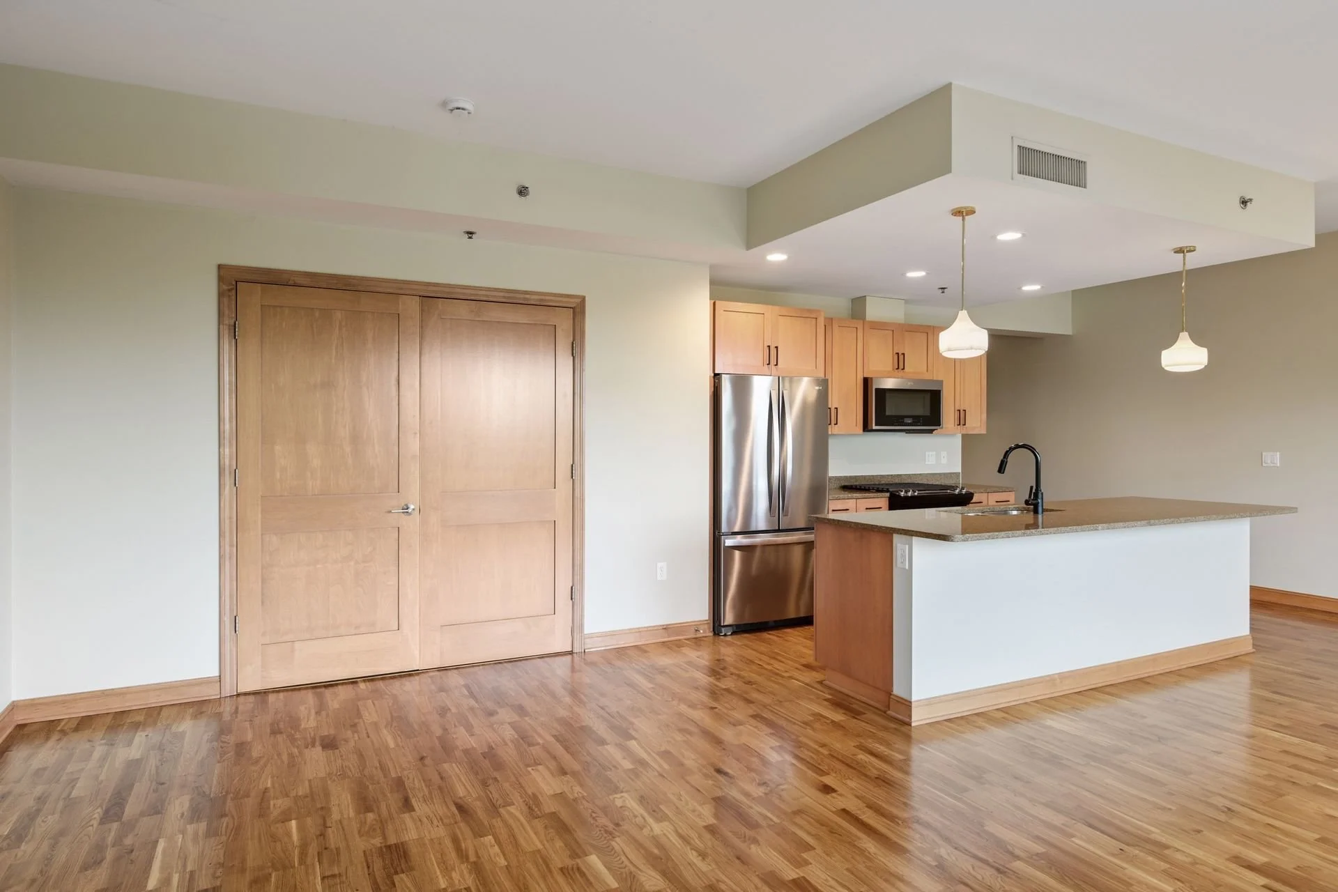Empty modern apartment with wooden floors, a kitchen with light wooden cabinets, stainless steel refrigerator, microwave, black sink and faucet, on a beige-colored wall with two hanging white pendant lights.
