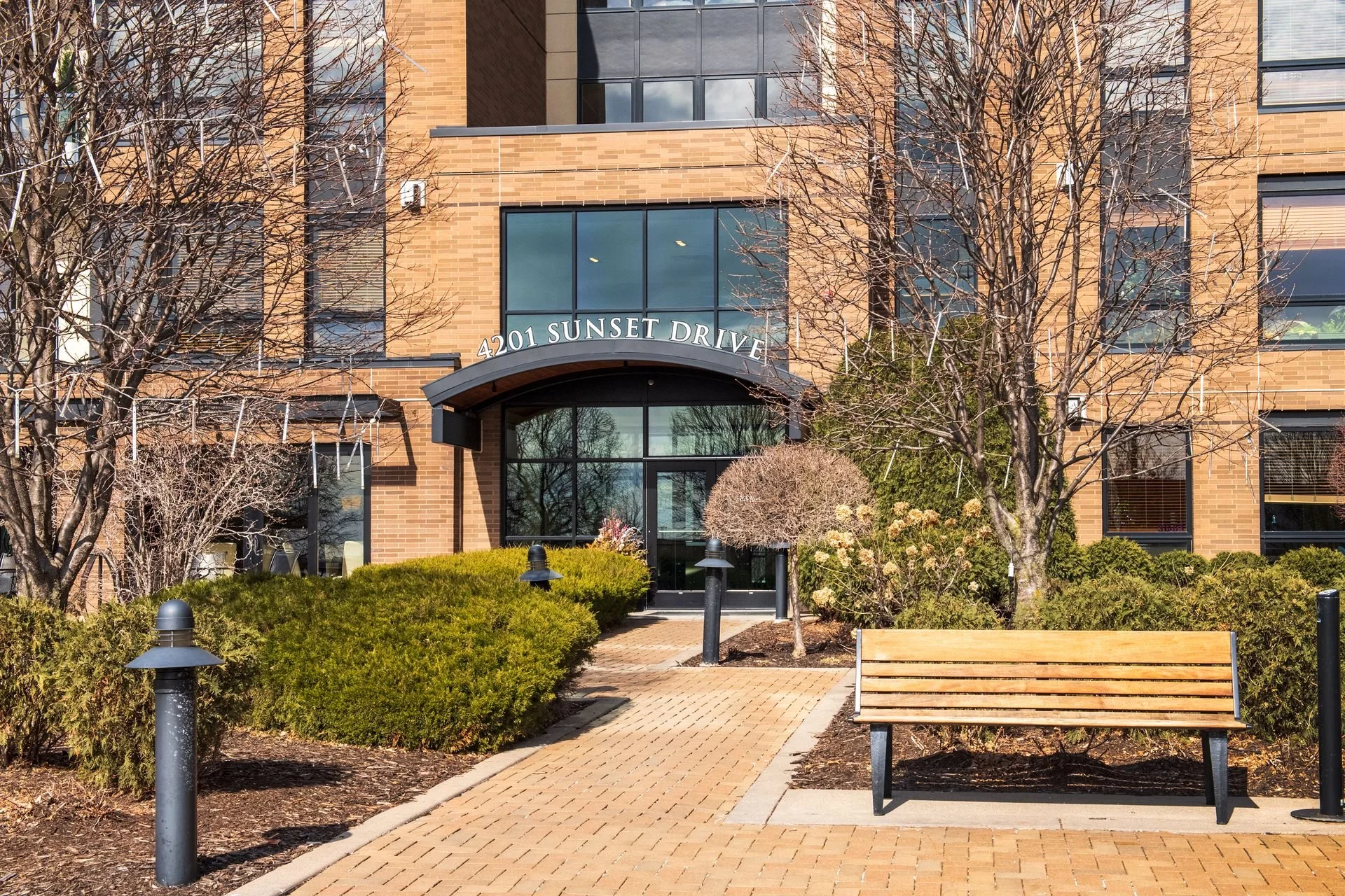 Apartment building entrance with a sign displaying '4201 SUNSET DRIVE'. There are trees, bushes, and a pathway leading to the entrance, with benches and outdoor lighting.