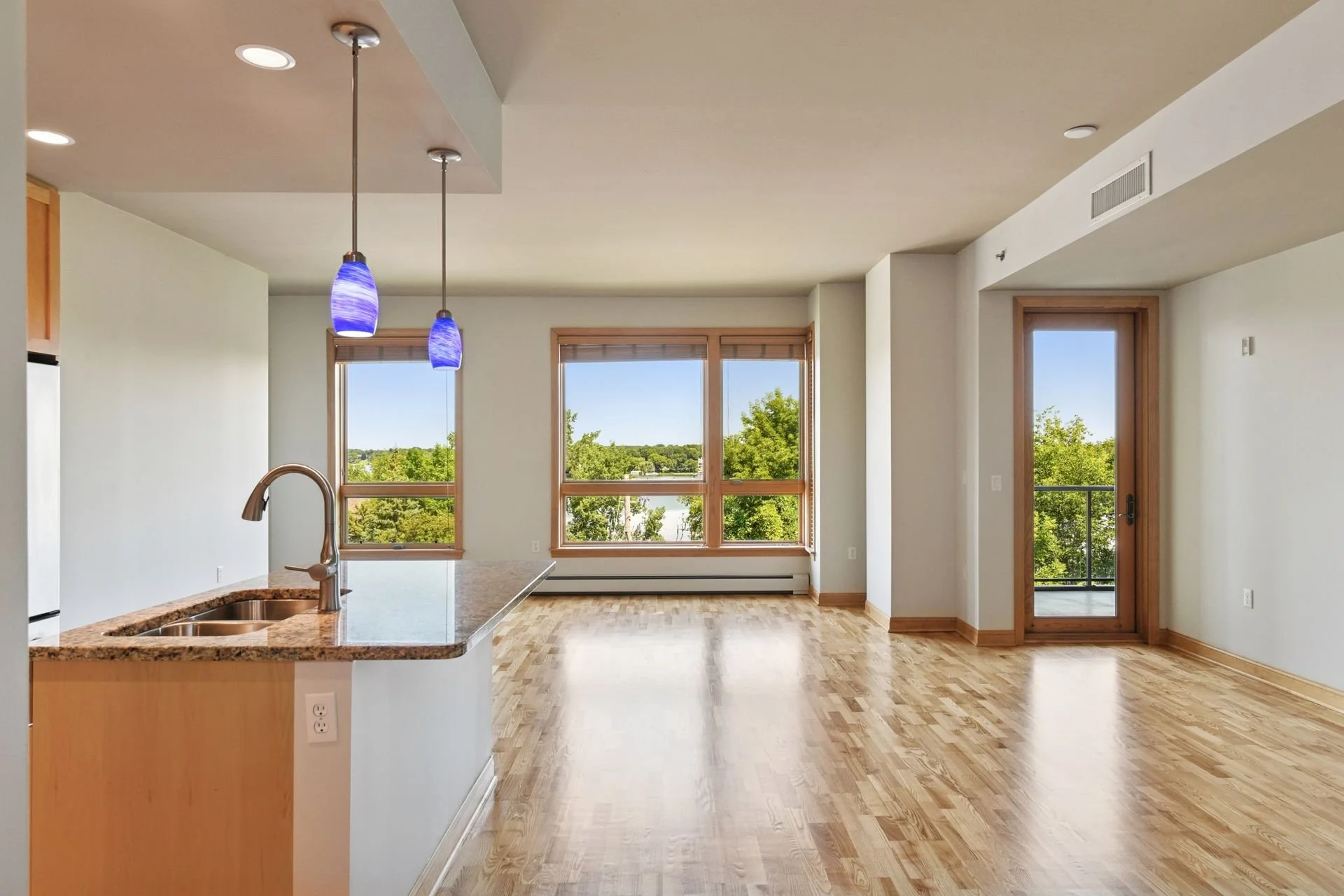 Living room with large windows showing trees outside, hardwood floors, a small balcony door, and a kitchen counter with blue pendant lights.