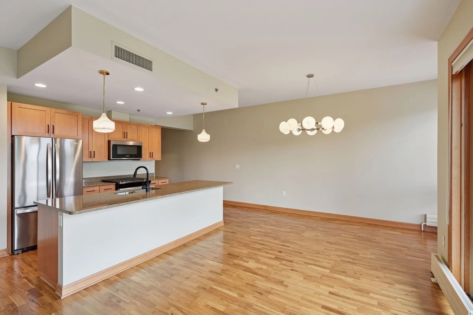Empty apartment with hardwood floors, kitchen with wooden cabinets, stainless steel refrigerator, microwave, and an island with a sink. Dining area with modern chandelier and pendant lights.