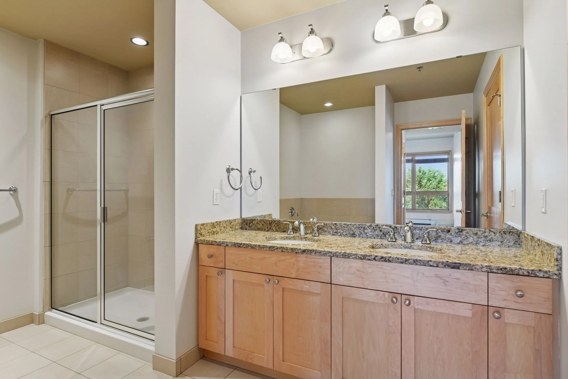 Bathroom with a corner shower, double vanity with granite countertop, large mirror, and window showing outdoor trees.