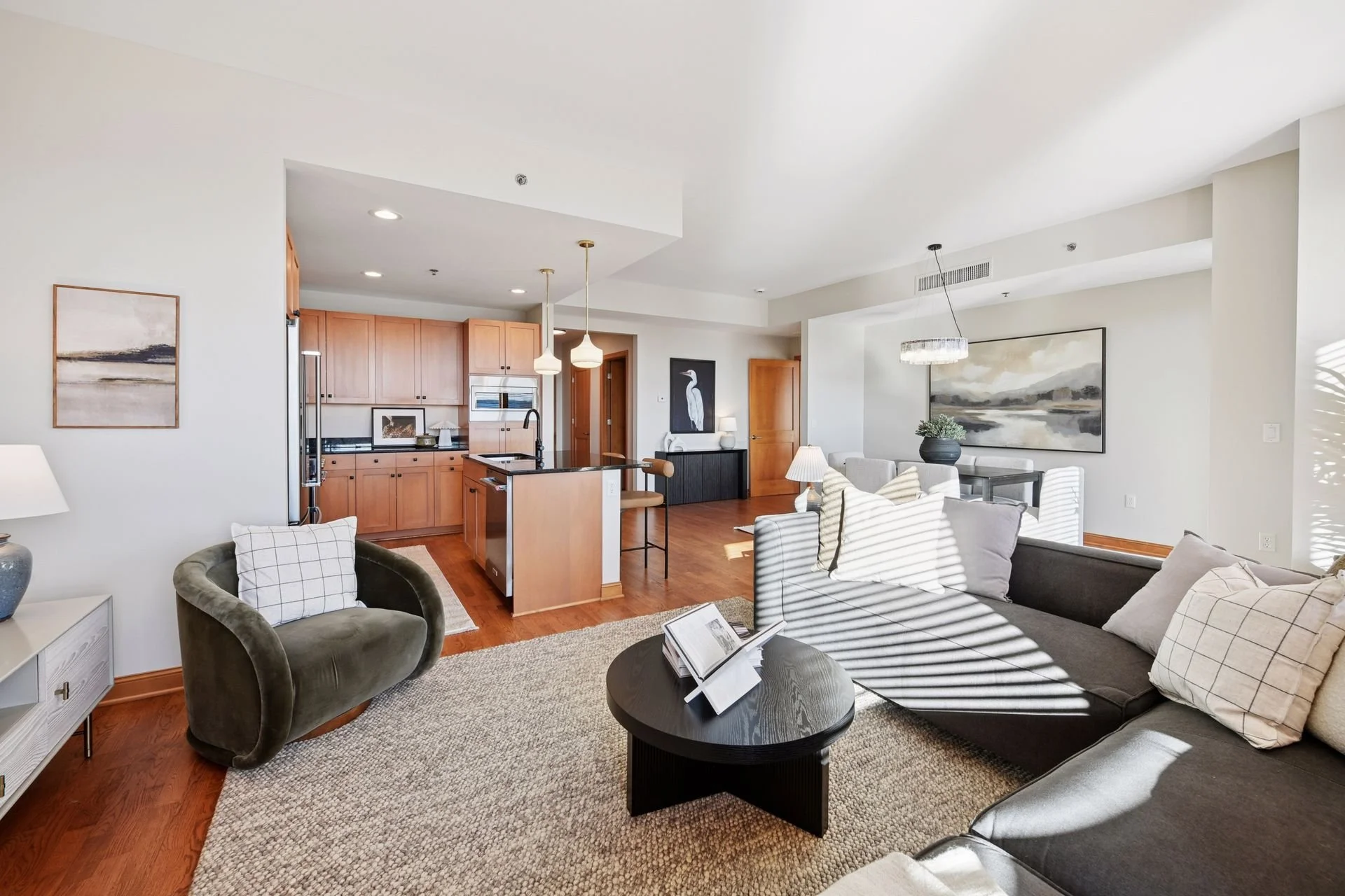 Open-concept living room and kitchen with wooden flooring and sunlight shining through window.