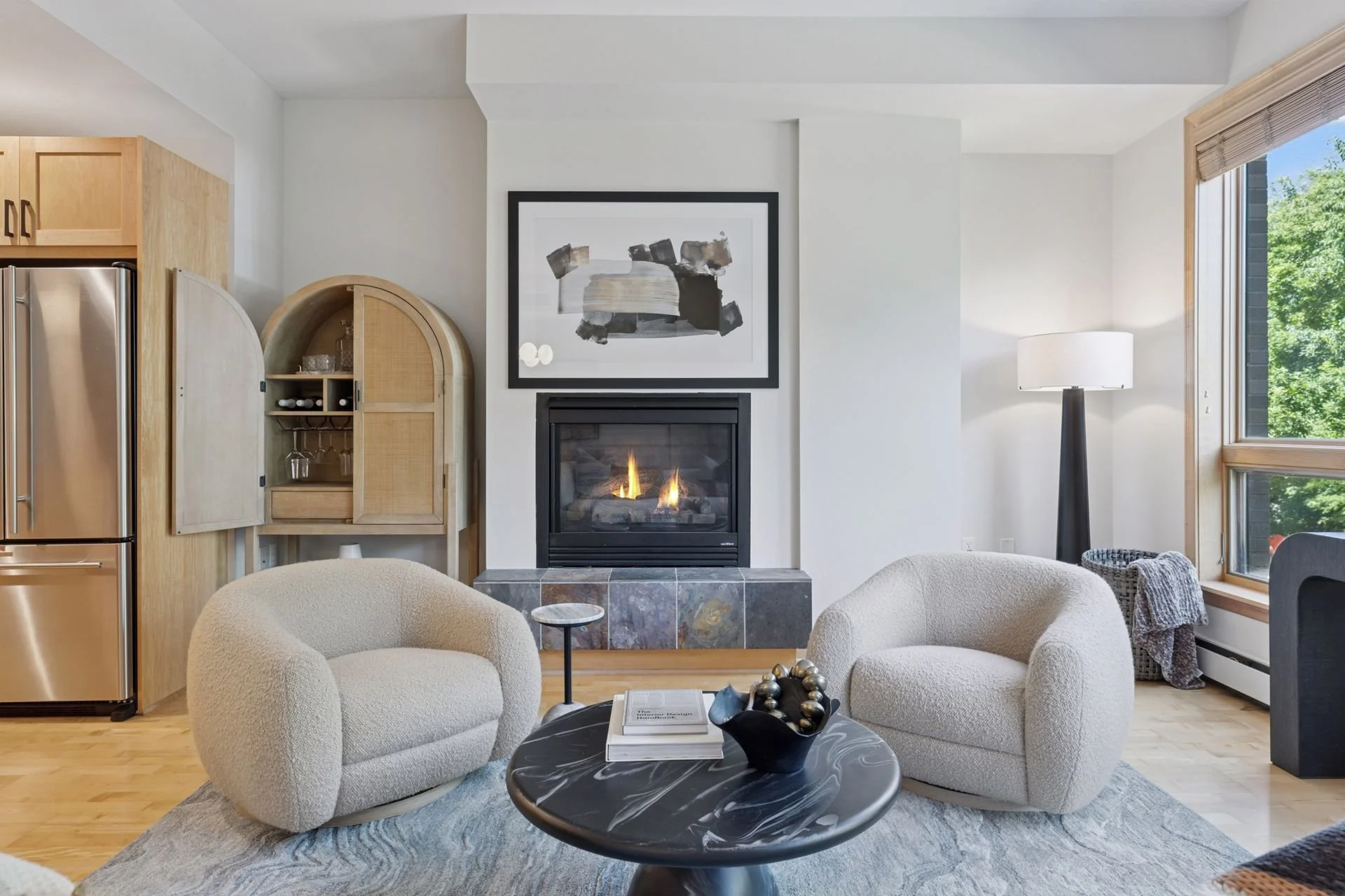 Living room with a gas fireplace with black frame, large window showing green trees, and wooden kitchen cabinetry to the left.