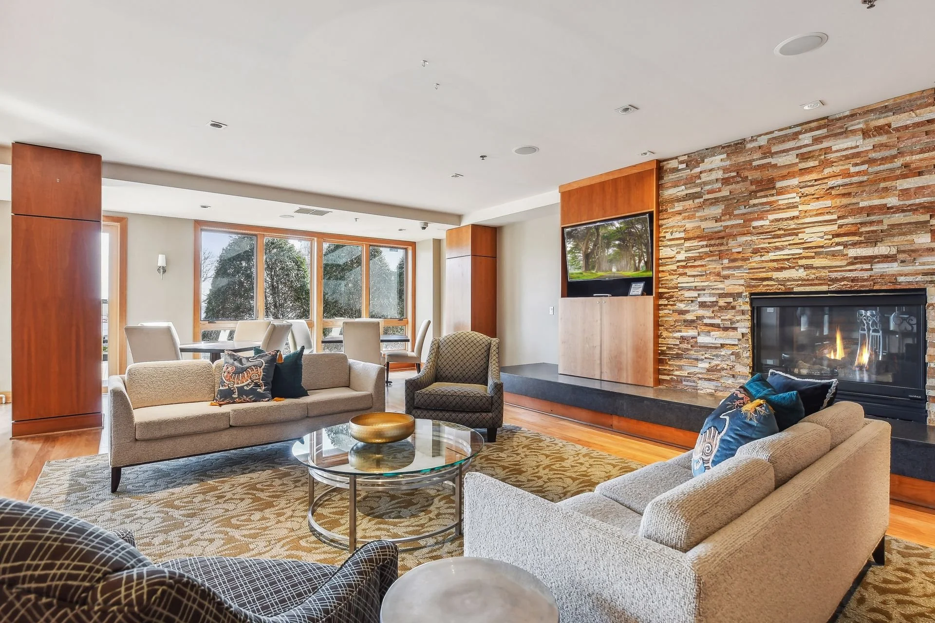 Cozy community room with sofas, a fireplace, a television, and large windows showing trees outside.