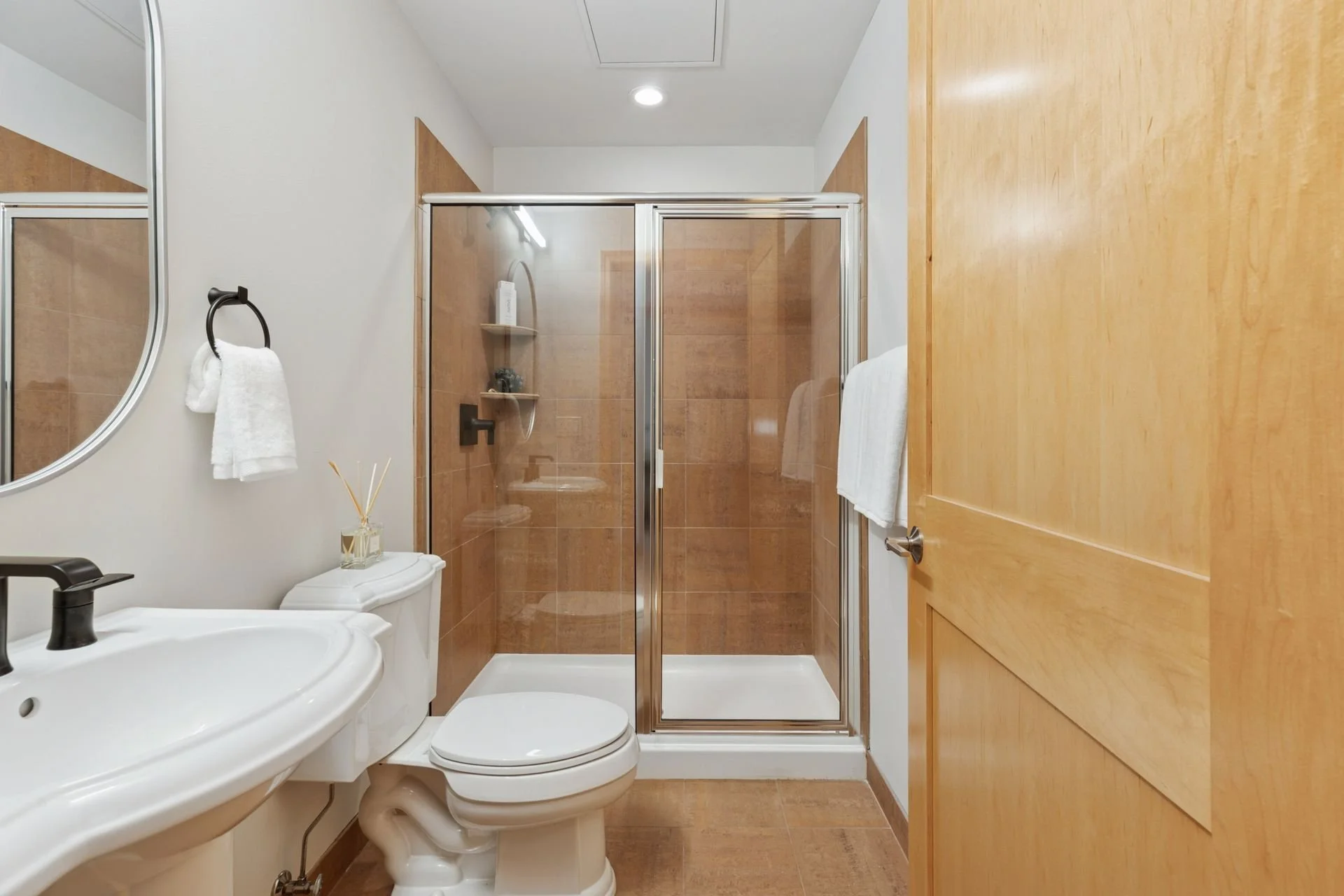 Bathroom with a shower, toilet, small sink, white towels, and light wood accents.