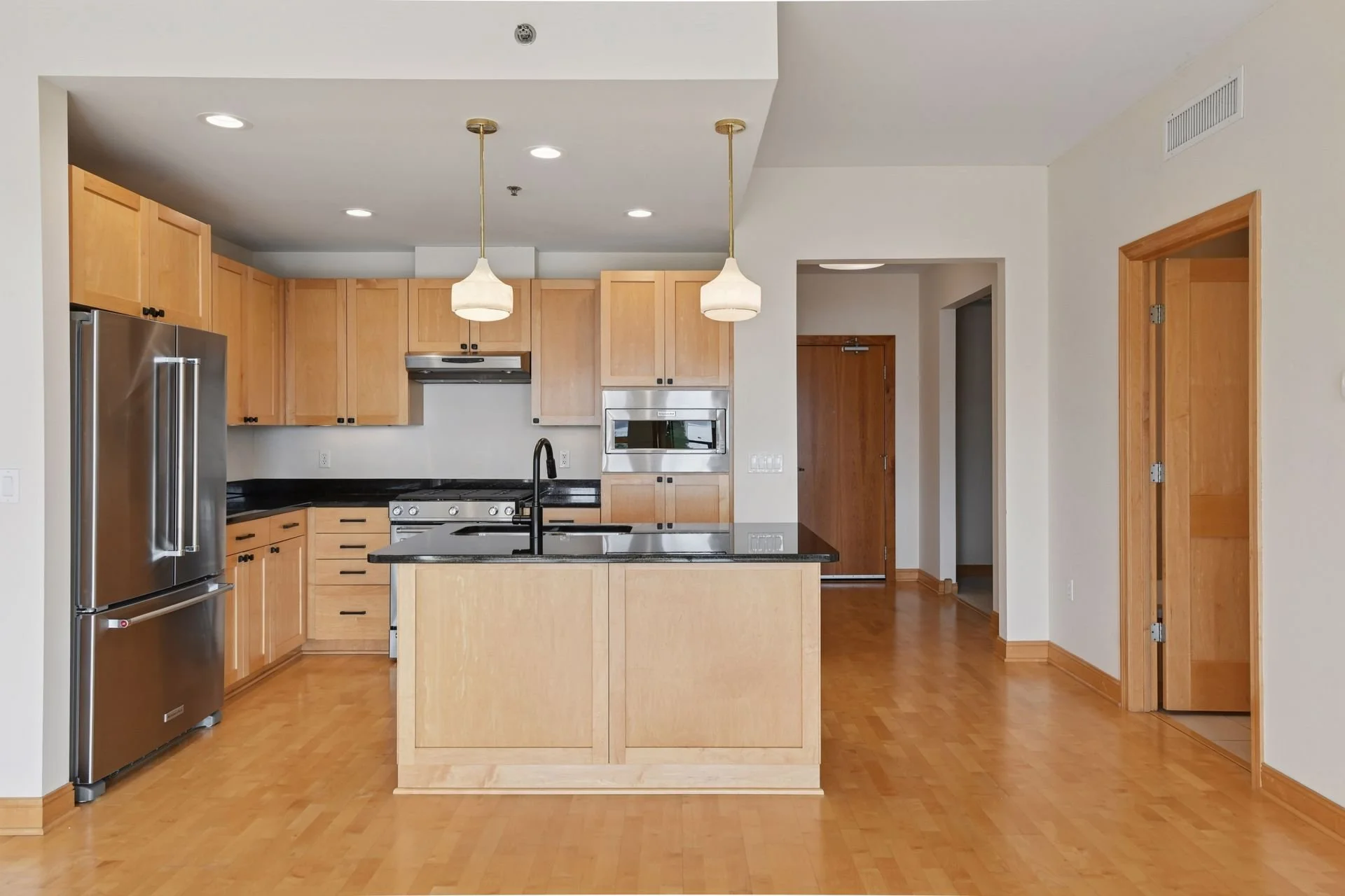 A modern kitchen with light wood cabinets, black countertops, stainless steel refrigerator, oven, and microwave. There are two pendant lights above a central kitchen island with a black faucet, hardwood floors, and multiple ceiling lights.