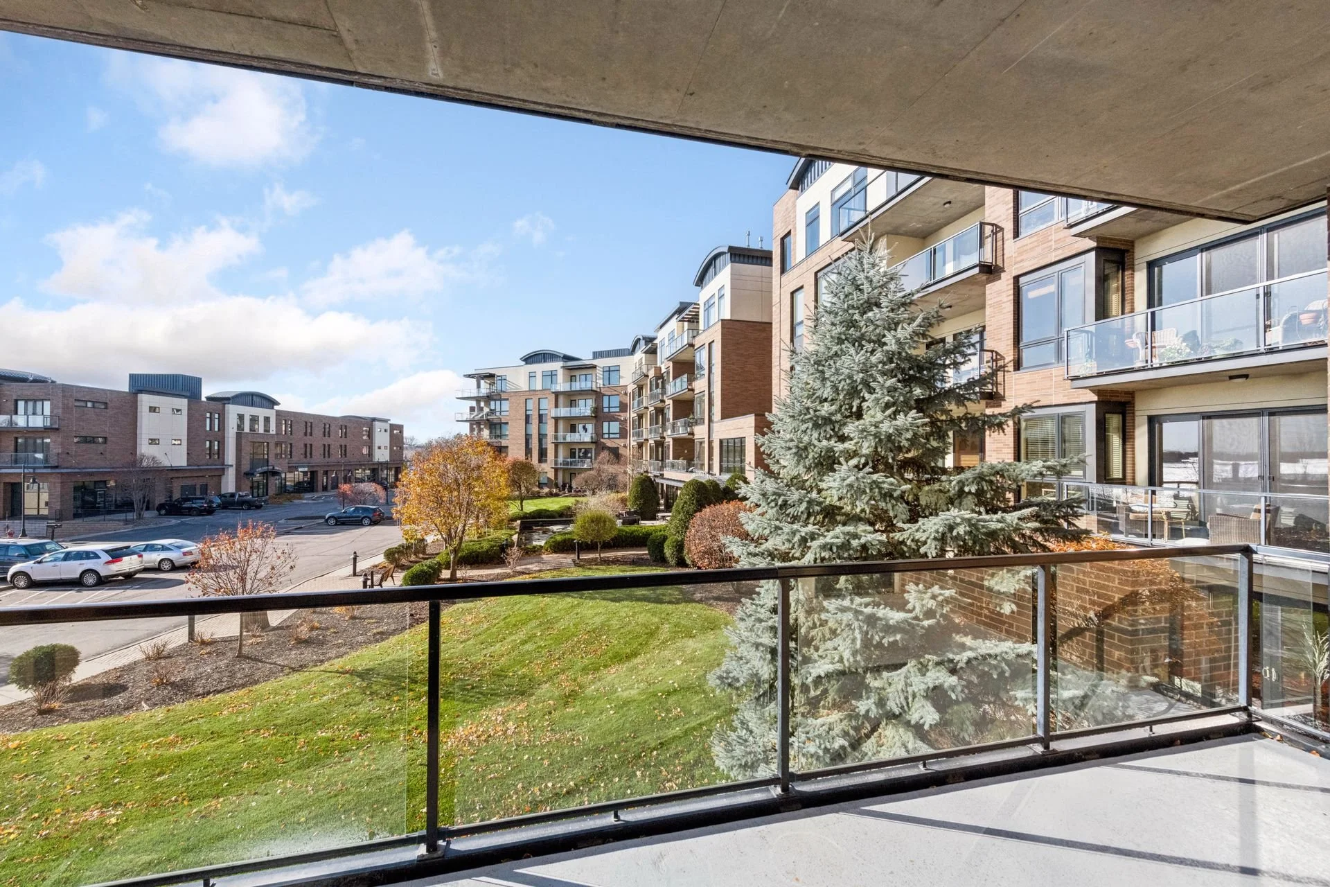 View from a balcony of a modern apartment building with trees, shrubs, and a parking lot under a partly cloudy sky.