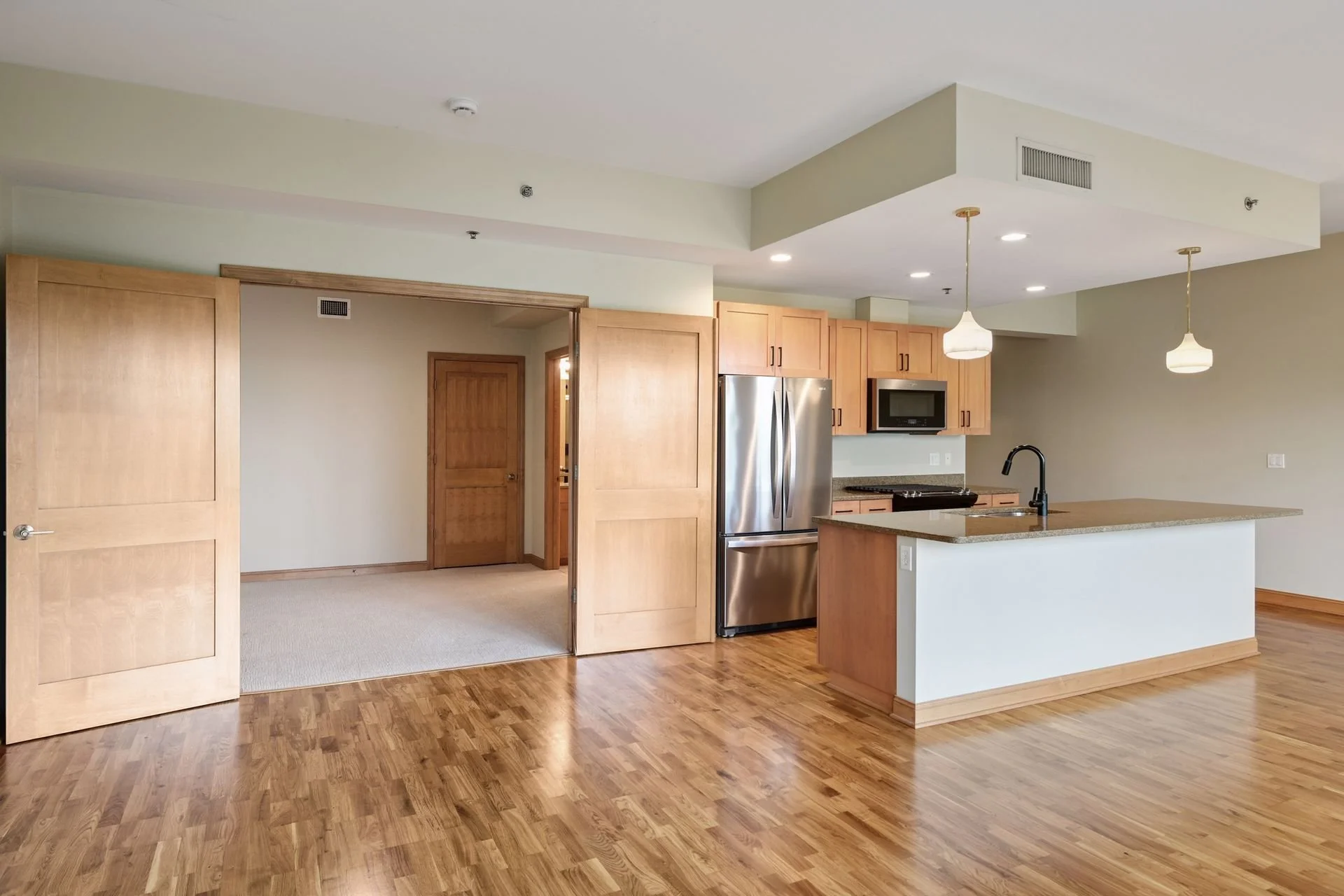 Open-concept kitchen with wooden cabinets, a stainless steel refrigerator, microwave, and island with a sink. Two pendant lights hang above the island, and there is a doorway leading to a carpeted room.