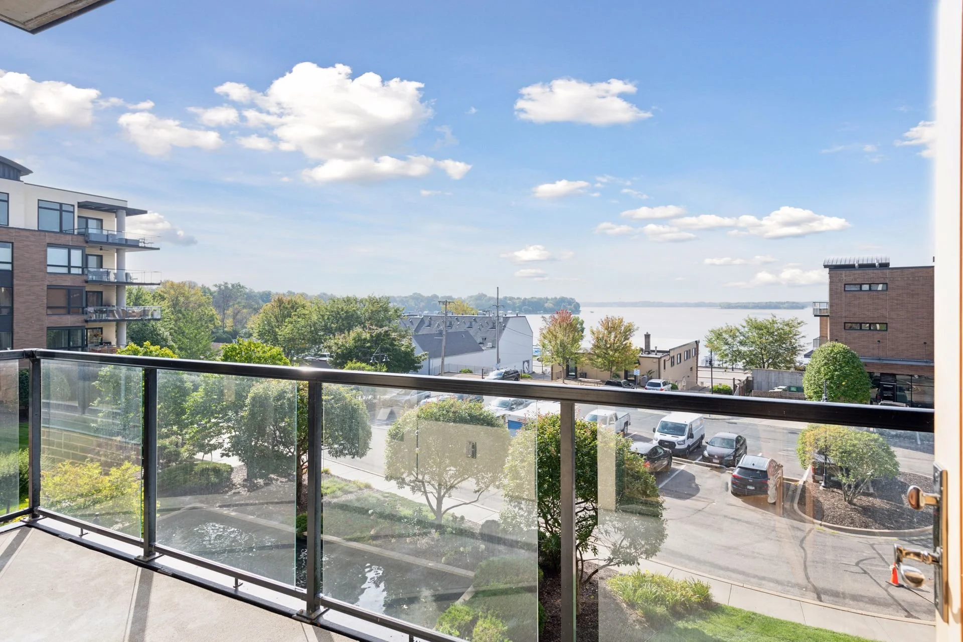 Balcony view overlooking a parking lot, trees, and buildings with water in the distance under a partly cloudy sky.