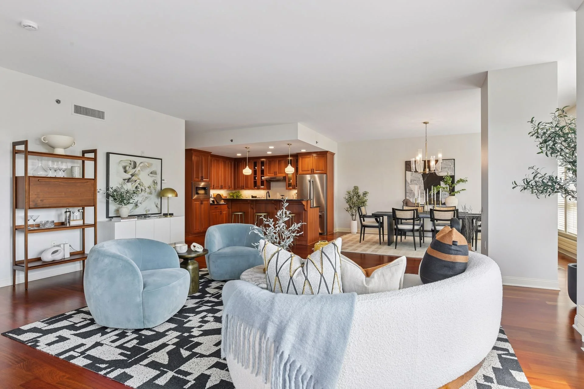Open-concept living room. In the background, a modern kitchen with wooden cabinetry, pendant lights, and stainless steel appliances are visible. To the right, a dining area with a chandelier.