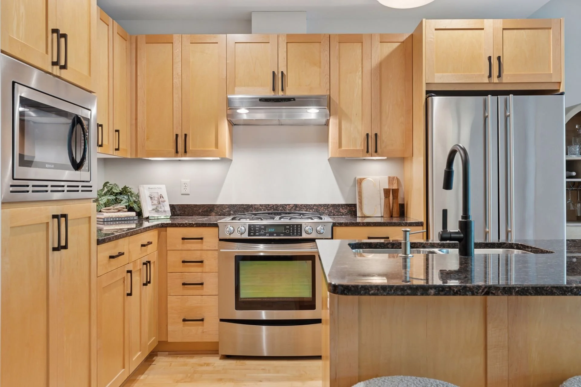 Modern kitchen with light wood cabinets, stainless steel appliances including a microwave, oven, and refrigerator. Black countertops, a black faucet, and a kitchen island are also visible.