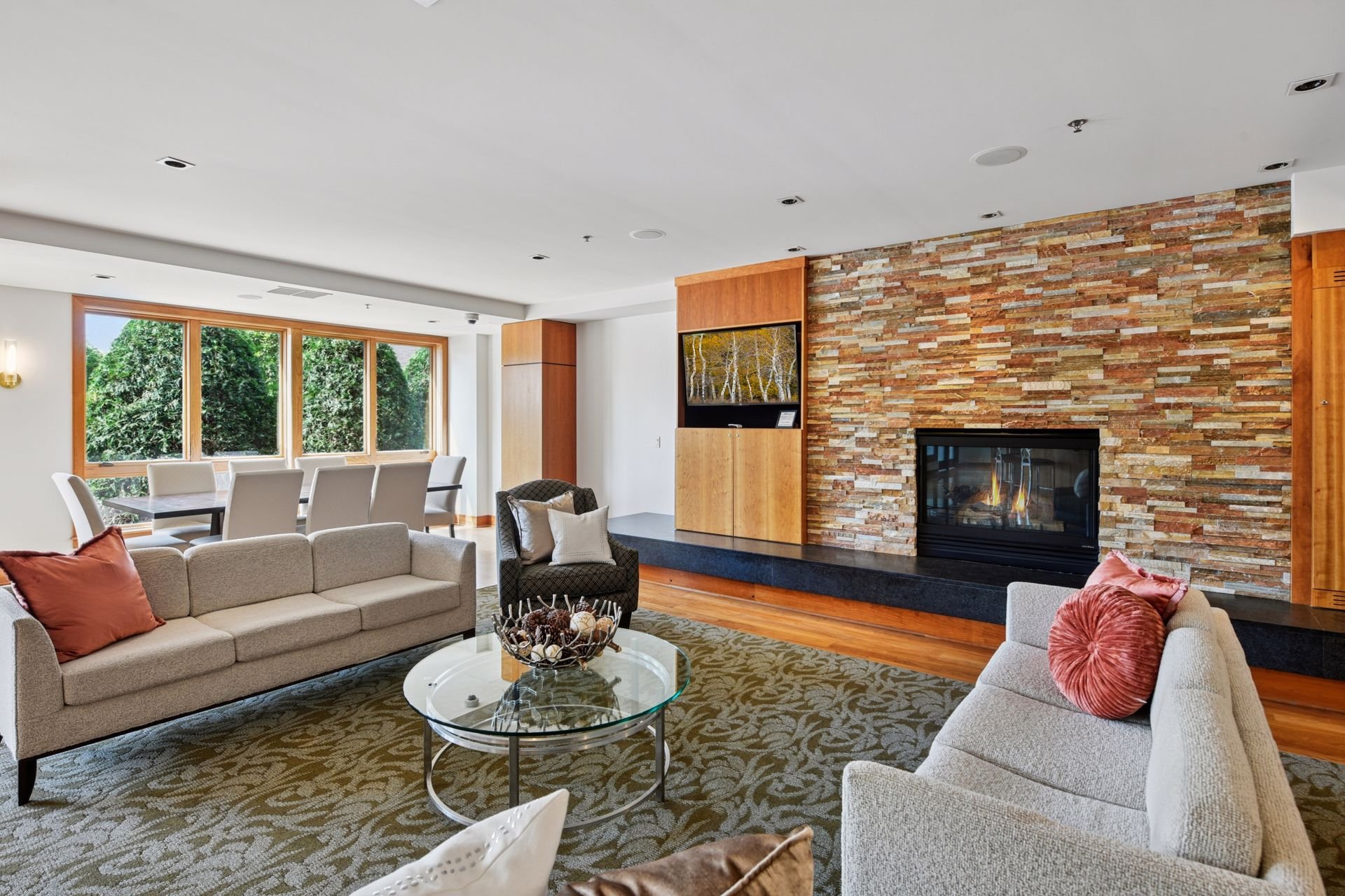 Community room with a fireplace, large windows with trees outside, and a dining area in the background.