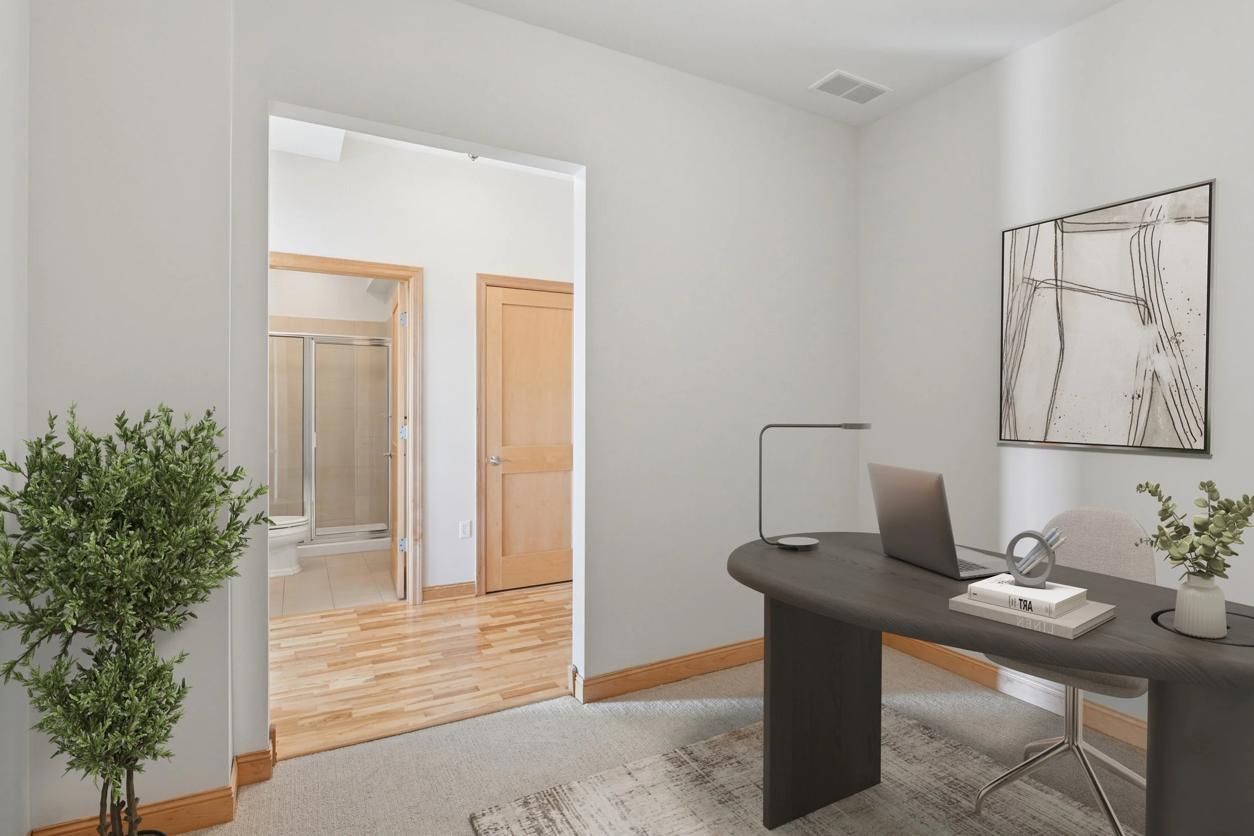 Home office with a dark wooden desk, a laptop, a table lamp, a gray office chair, and a white vase with greenery, with a framed abstract art piece on the wall, and a view into a bathroom with a glass shower door.