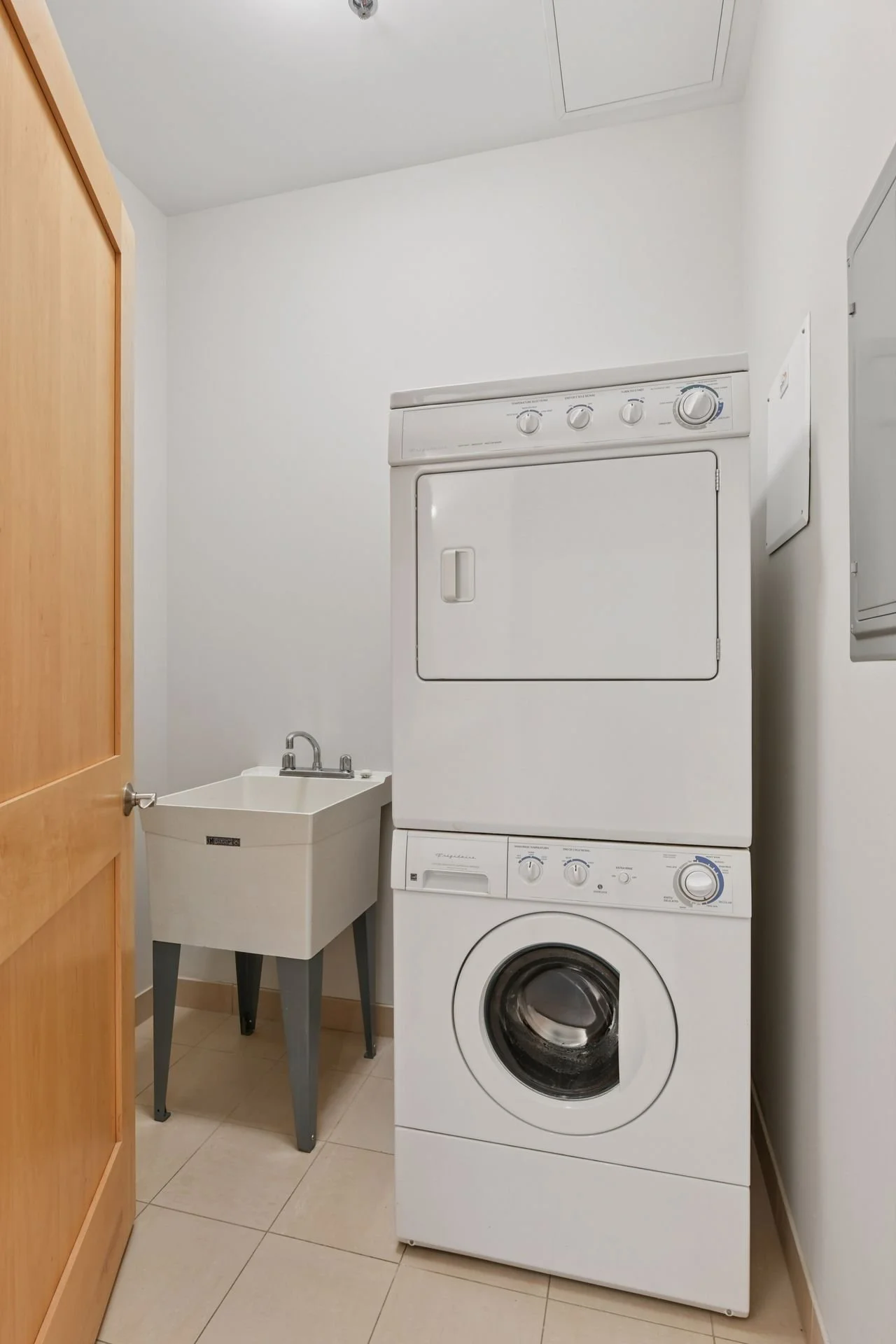 A laundry room with a stacked washer and dryer, a small utility sink, and beige floor tiles.