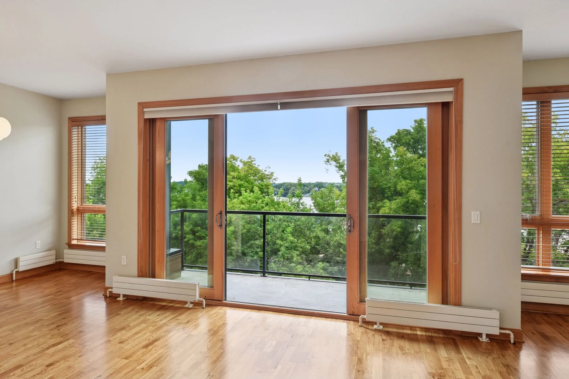 Living room with wooden floors, large sliding glass doors leading to a balcony, and windows with wooden blinds, overlooking green trees.