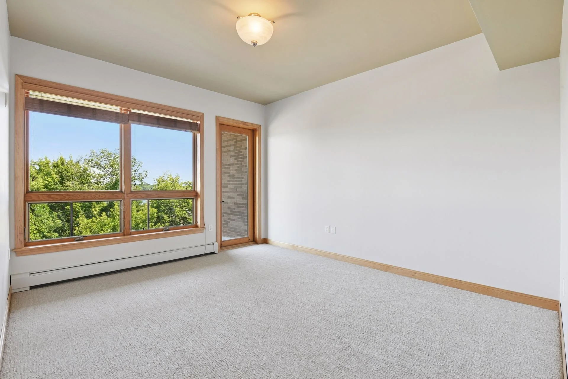 Large bedroom with large window and door, beige carpet, white walls, wooden trim, and greenery outside