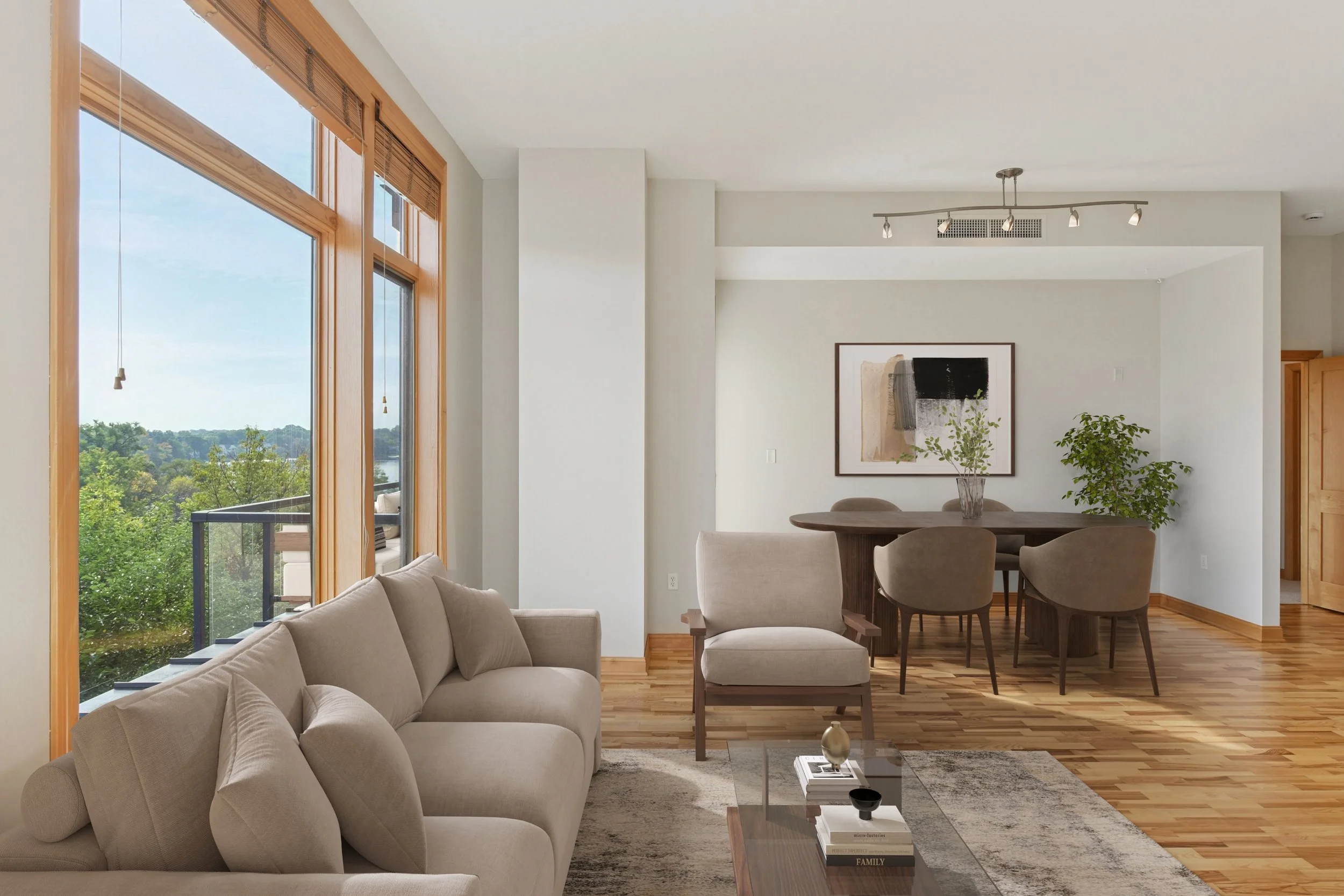 Living room with beige sofa and armchair, wooden floors, large windows with wooden frames overlooking trees and sky, round dining table with chairs, abstract art on wall, potted plants, coffee table with books and decorative objects.