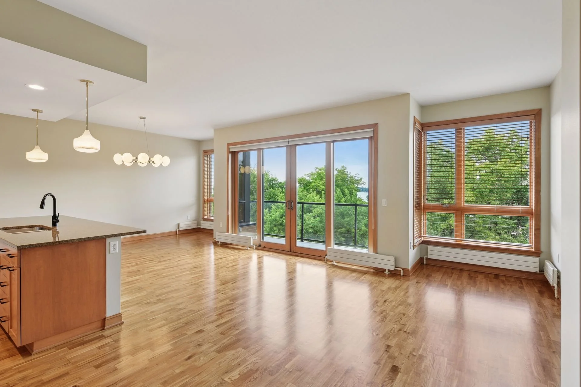 Empty living room with hardwood floors, large windows with wooden blinds, and a balcony door, in a modern apartment.