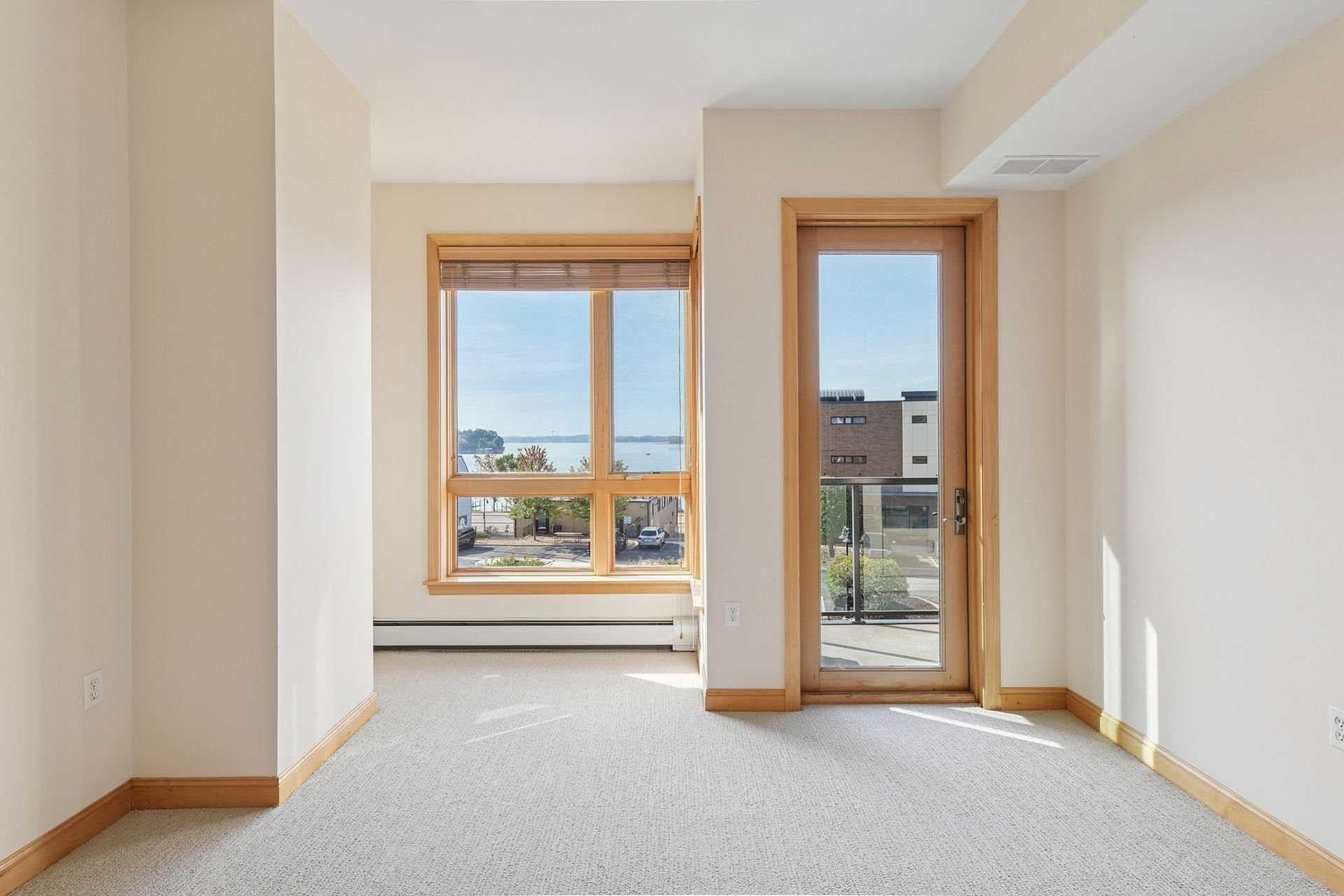 Empty room with large window and glass door showing a view of the street and water, beige walls, light-colored carpet, and wood trim and baseboards.